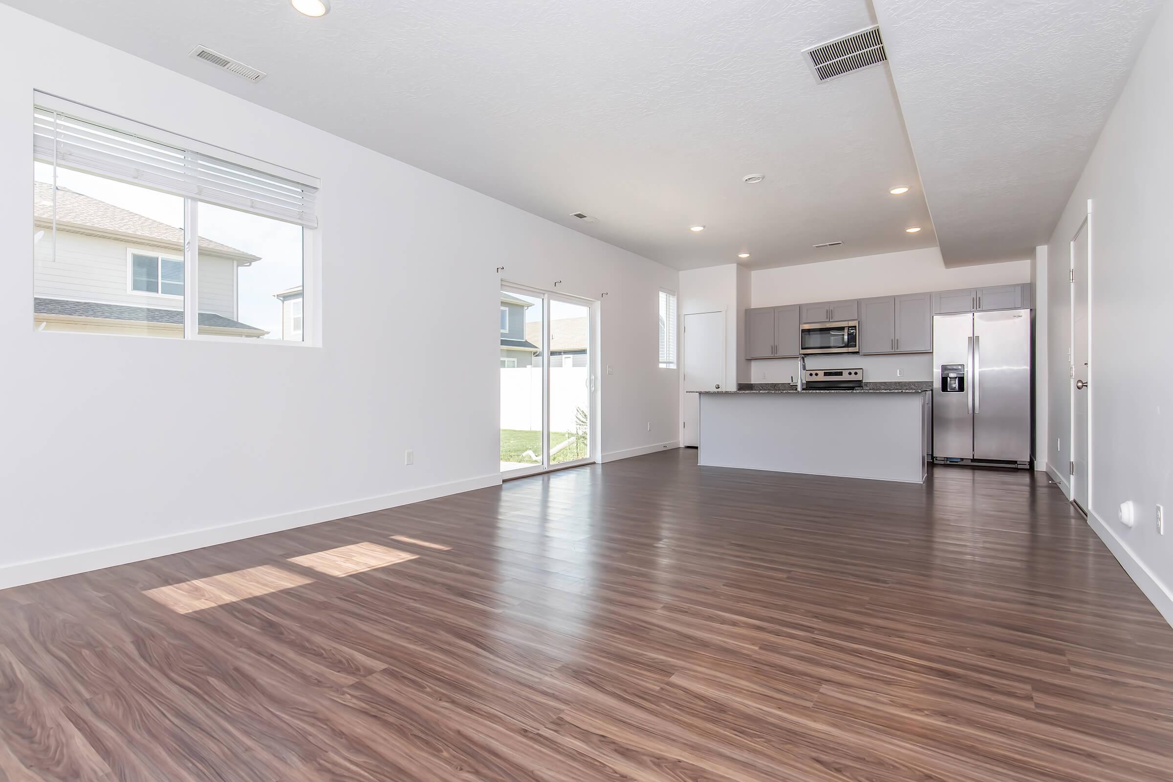 A bright, modern living space featuring wood laminate flooring, large windows, and an open layout. The kitchen is equipped with stainless steel appliances and gray cabinetry. Sliding glass doors lead to an outdoor area, illuminating the room with natural light.