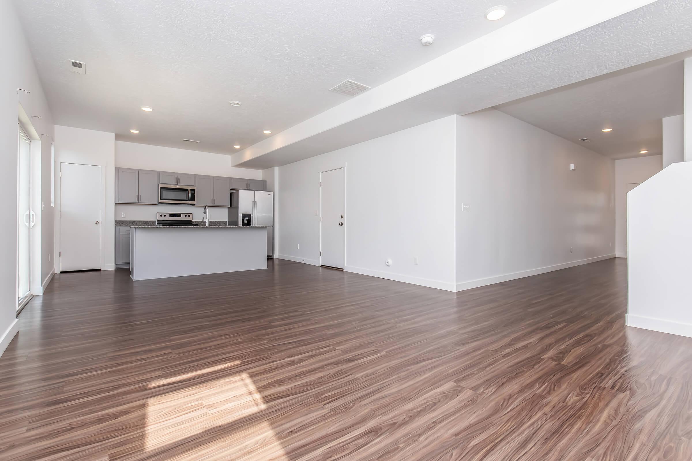 A spacious, modern open-concept living area with hardwood flooring. The kitchen area, featuring stainless steel appliances and gray cabinetry, is partially visible. Large windows allow natural light to fill the room, which has white walls and ceiling lights. The layout is bright and inviting.