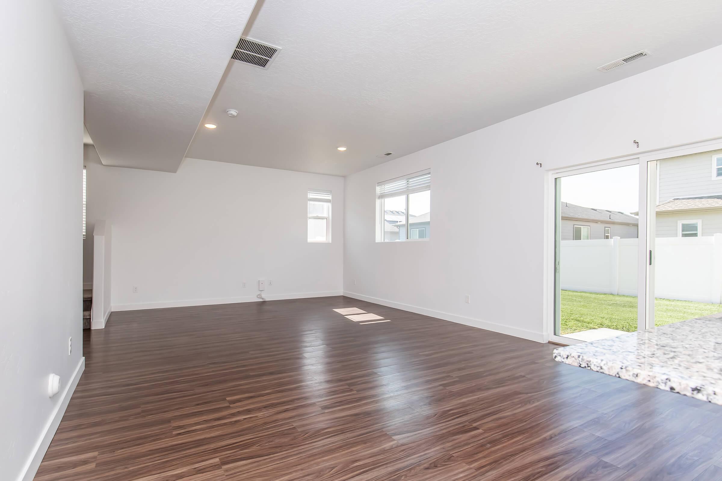 Spacious, empty living room with light-colored walls and hardwood flooring. Two windows allow natural light to fill the space, and a sliding glass door opens to a backyard. The kitchen area features a granite countertop. The overall atmosphere is bright and modern, perfect for decorating.
