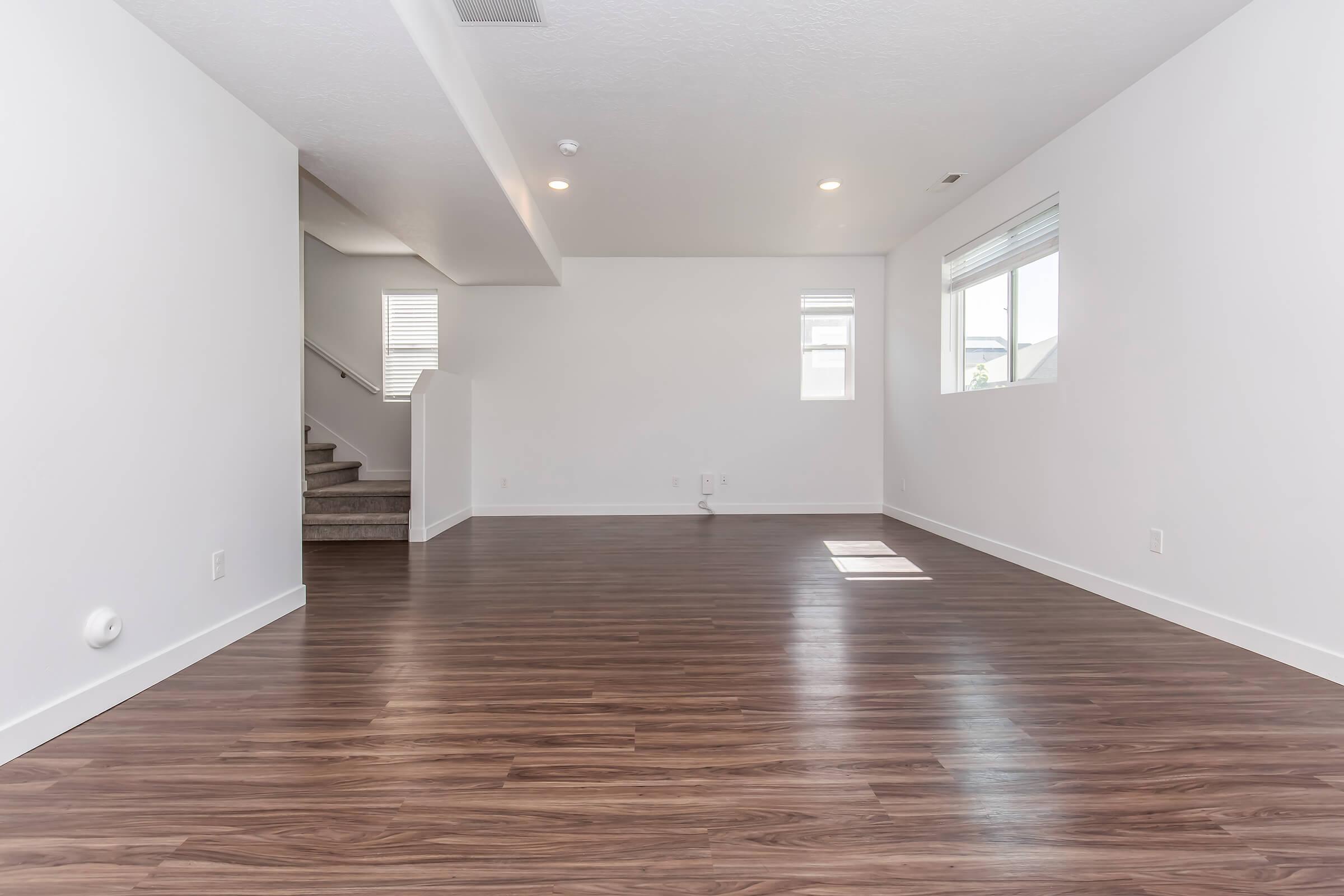Empty modern living room with wood laminate flooring, featuring a large window allowing natural light, white walls, and a staircase in the background. The space is uncluttered, creating a bright and open atmosphere.