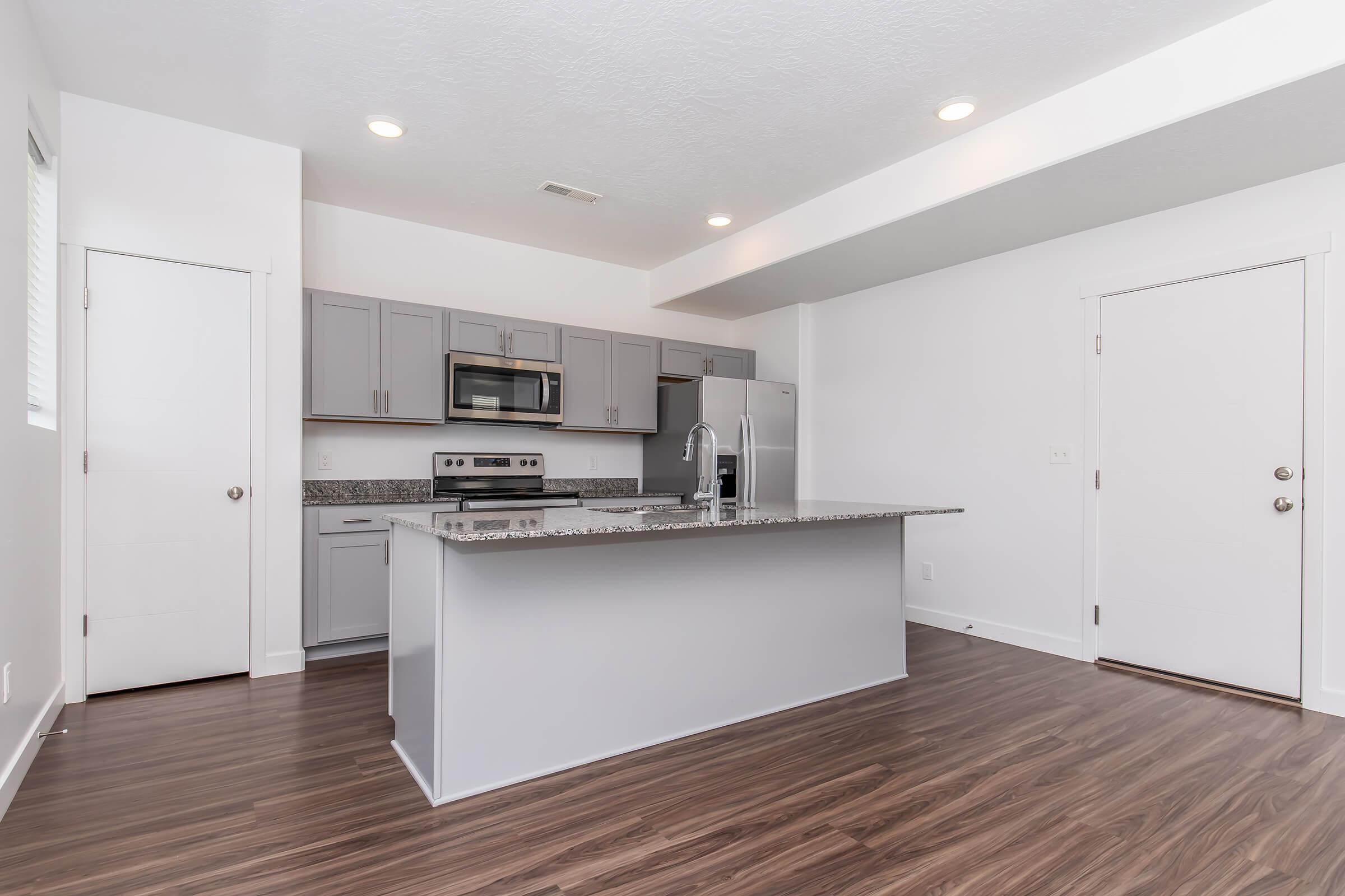 Modern kitchen featuring gray cabinets, stainless steel appliances, and a granite countertop with a large island. The space has dark wood flooring and ample natural light from a nearby window, creating a bright and inviting atmosphere. Simple design emphasizes functionality and style.