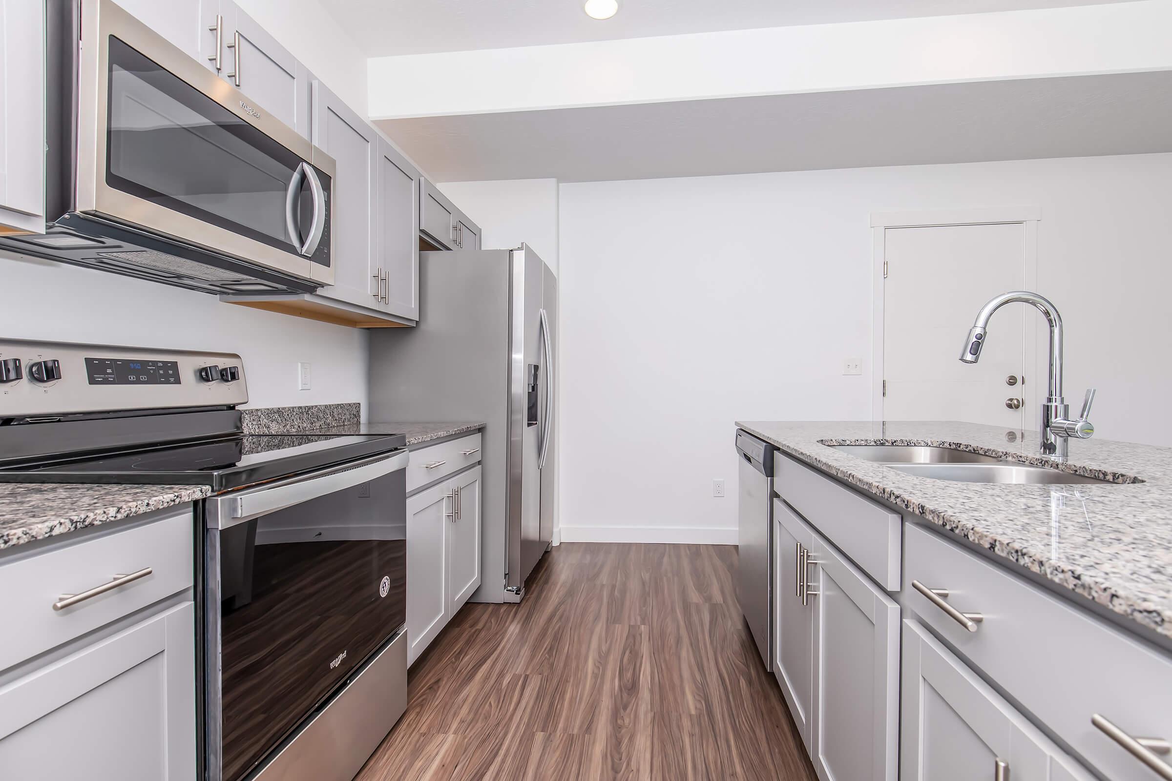 Modern kitchen featuring stainless steel appliances, including a microwave, oven, and refrigerator. The countertops are made of gray granite, and the cabinetry is light gray. The open layout includes a view of glossy wooden flooring and ample natural light from nearby windows.