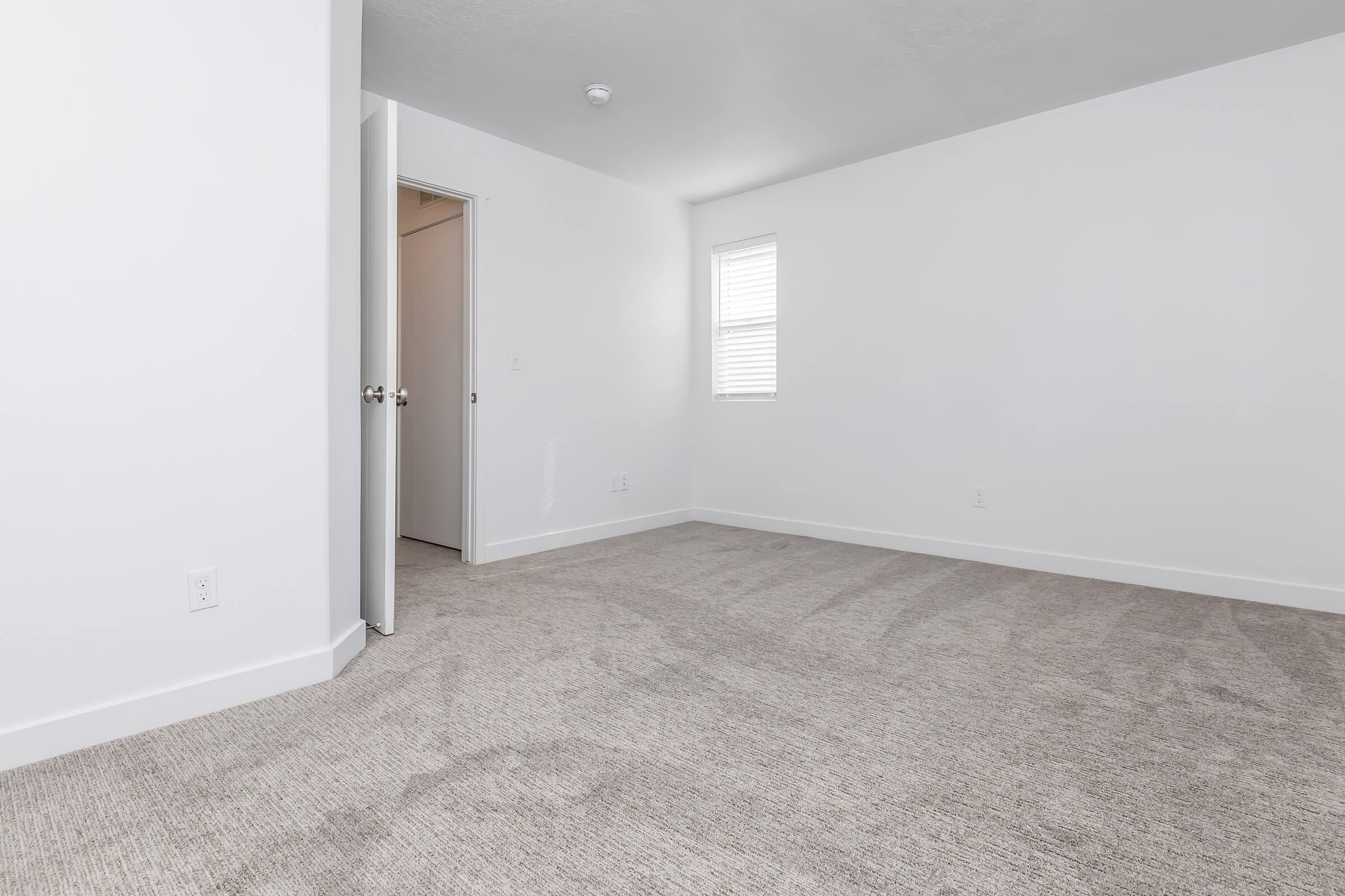 A clean, empty room with light gray carpet and white walls. There's a partially open door on the left leading to another area, and a small window with blinds on the right allowing natural light to fill the space. The room is bright and spacious, ideal for various purposes.