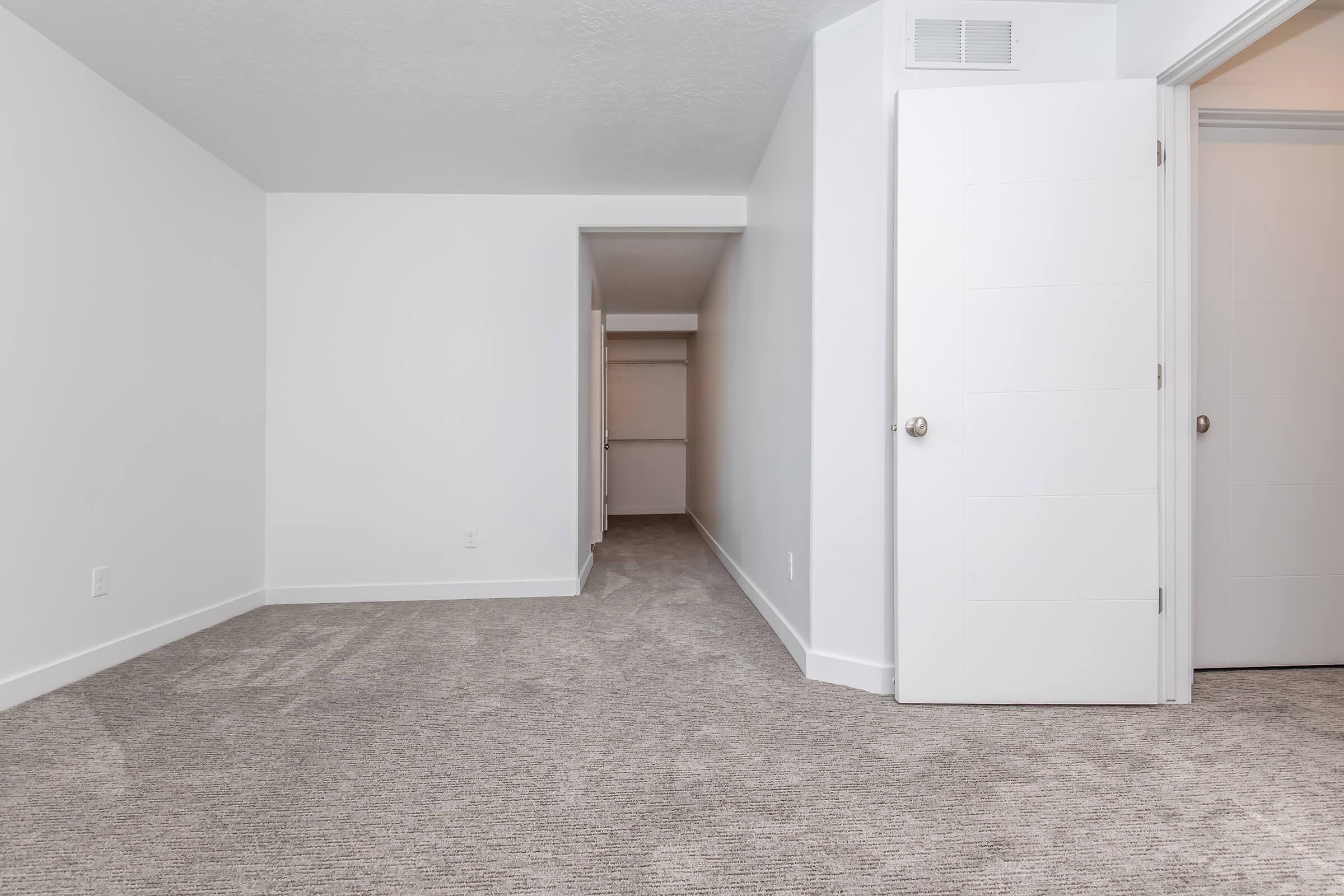 A well-lit room with light gray carpeting and white walls. A doorway leads to a small hallway with an open closet on the right. A white door is partially open to the right, suggesting access to another space. The room appears spacious and clean, suitable for various interior design options.