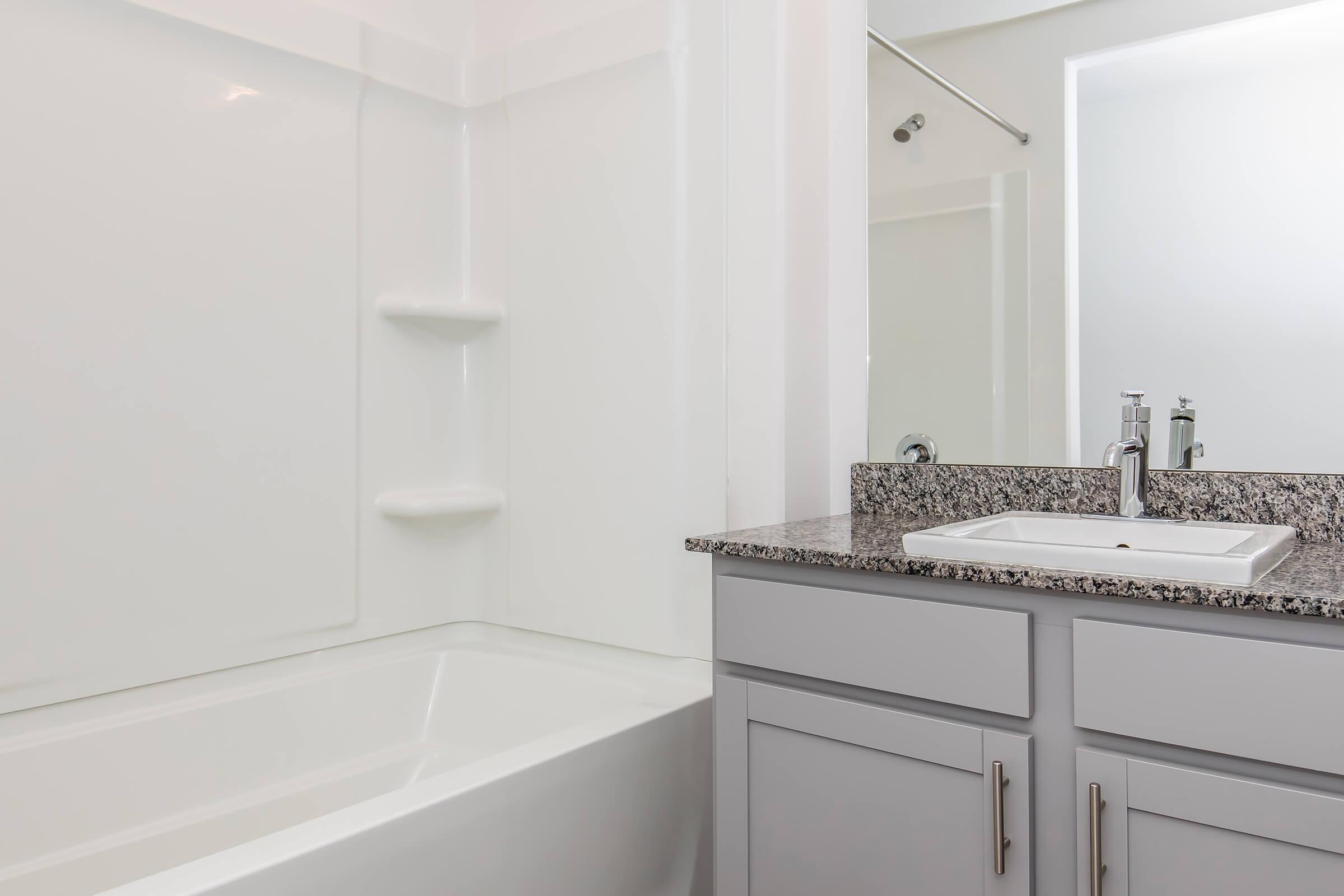 A modern bathroom featuring a white bathtub and shower combination on the left. A grey vanity with a granite countertop and a rectangular sink is positioned on the right. The space is well-lit with neutral-colored walls, creating a clean and contemporary atmosphere.
