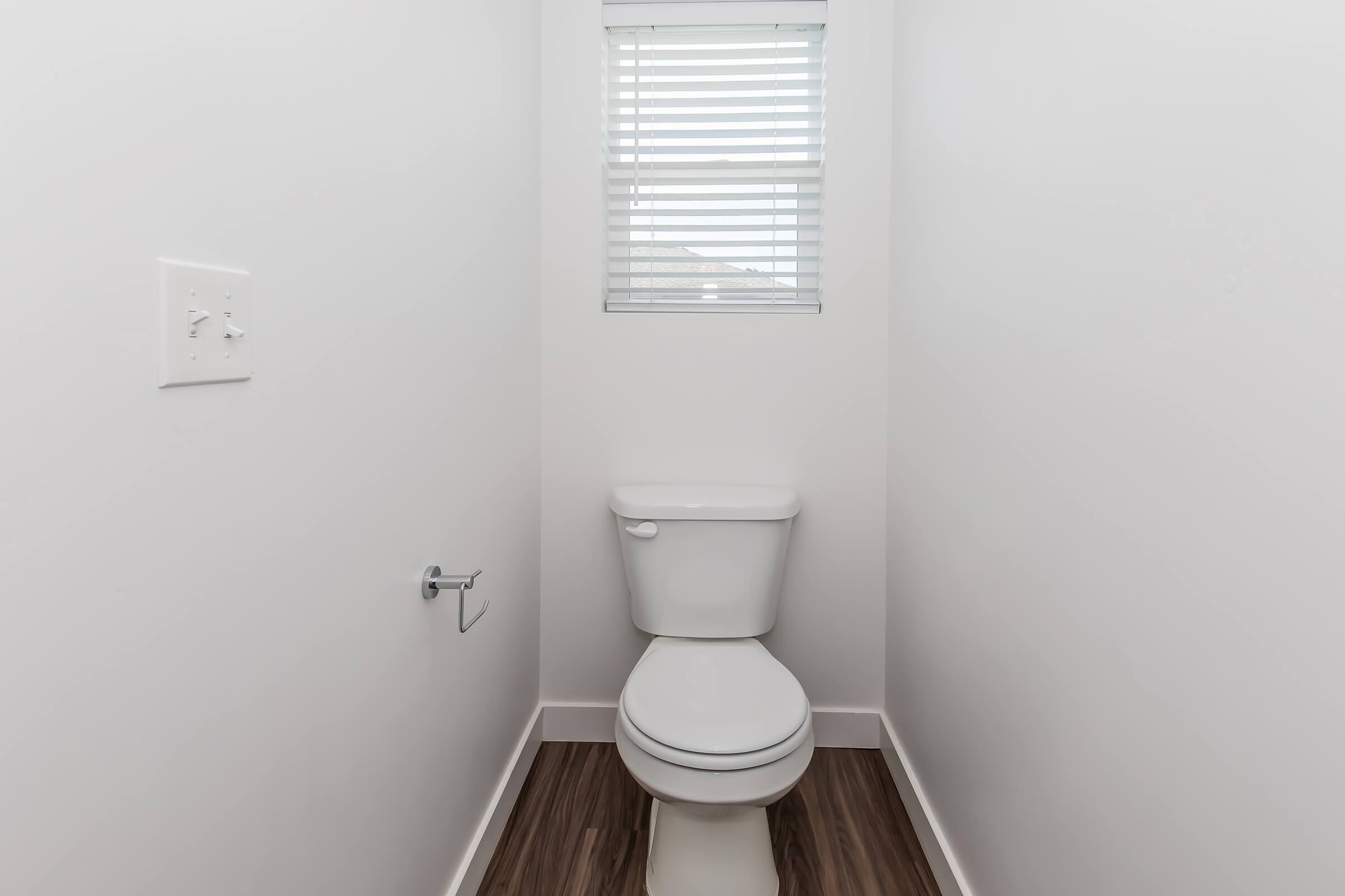 A minimalist bathroom with a white toilet, a toilet paper holder on the wall, and a small window featuring blinds. The walls are plain and white, and the floor has a wooden appearance, creating a clean and simple aesthetic.
