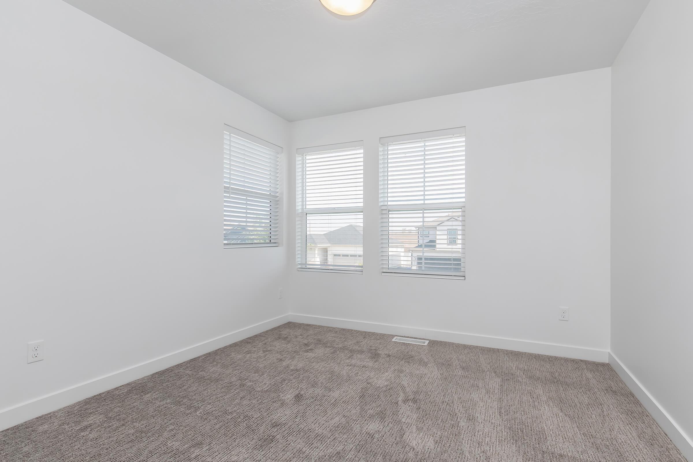 A clean, empty room with light gray carpeting and white walls. Three windows with blinds allow natural light to enter, providing a bright atmosphere. The space is unfurnished and has a simple ceiling light fixture.