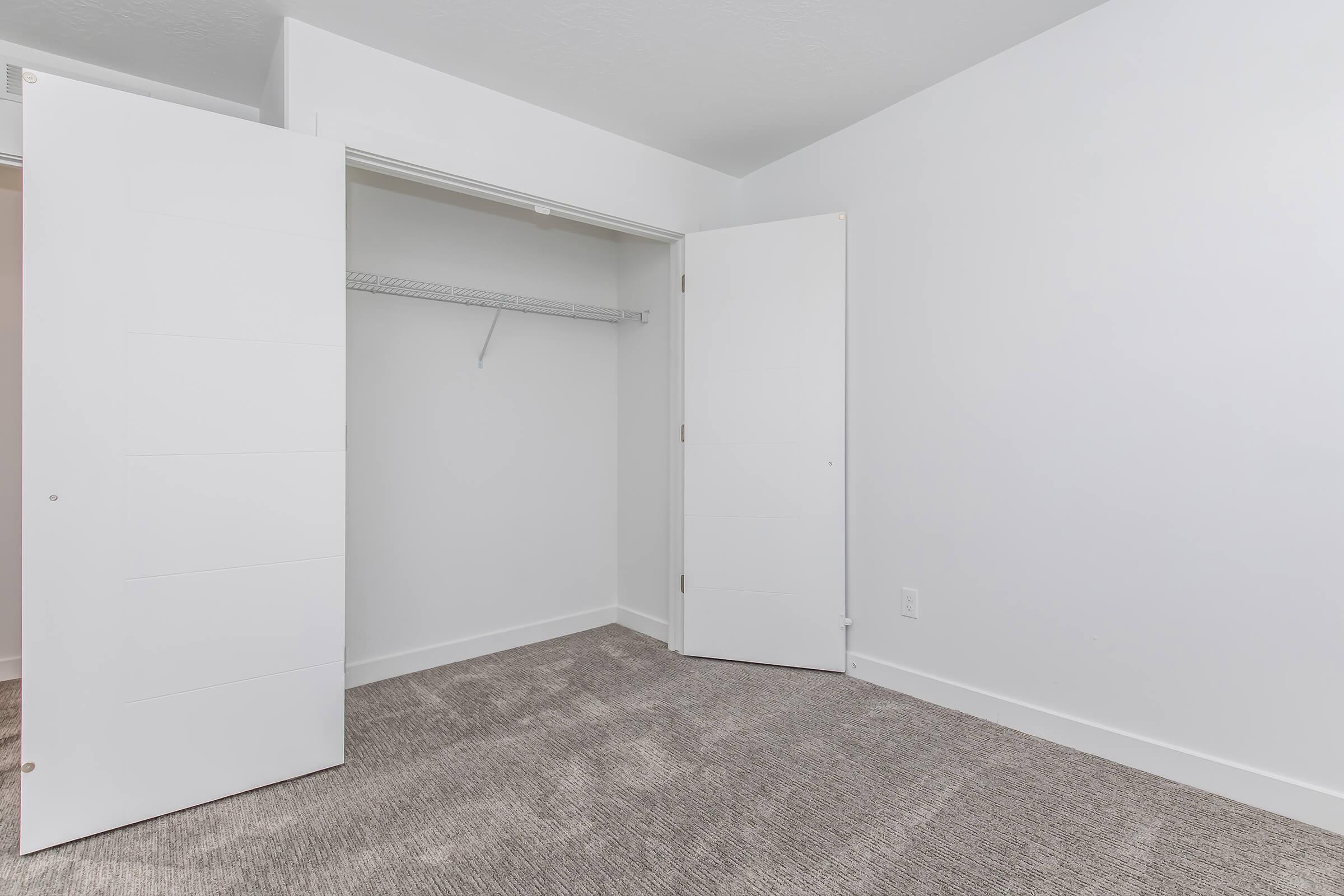 Empty room with beige carpet and white walls. An open closet with a metal wire shelf is visible, featuring two white doors that swing outward. Natural light illuminates the space, giving it a clean and spacious appearance.
