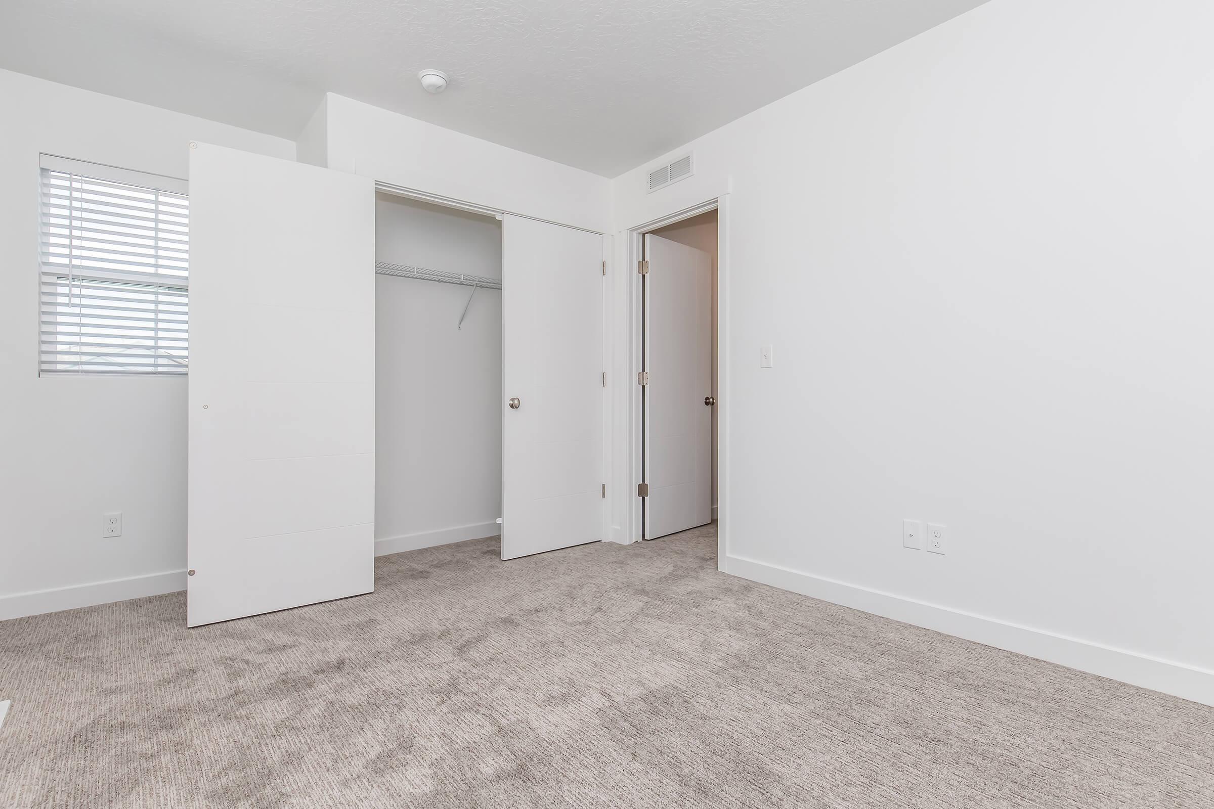 A light-colored room with soft beige carpet, featuring two white doors that lead to a closet and another room. A window with blinds allows natural light in, and the walls are painted white, giving the space a clean and airy feel.