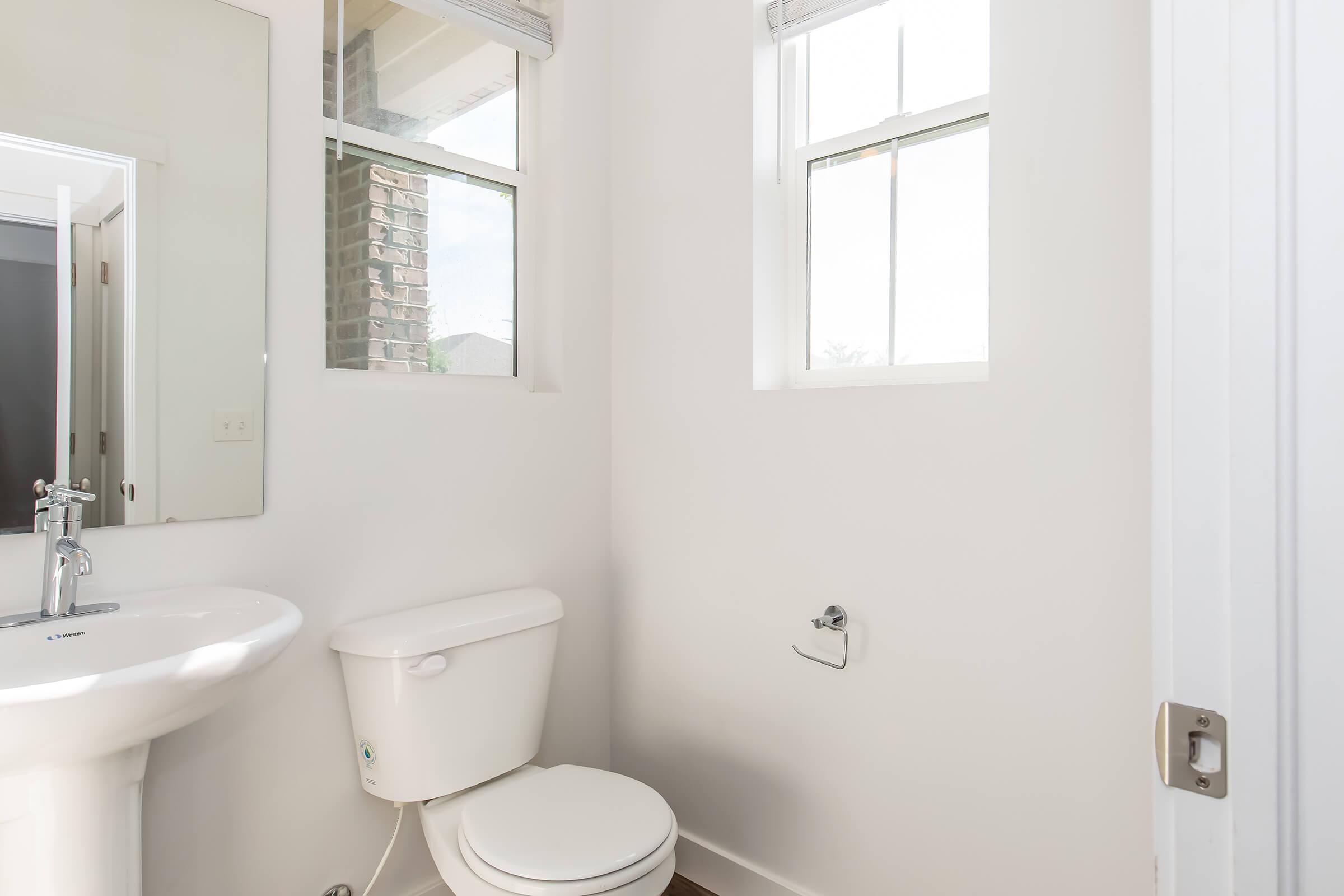 A clean, modern bathroom featuring a white toilet, a sink with a mirror above it, and two windows allowing natural light. The walls are painted white, and the floor is a dark material. A door is partially visible on the right side.