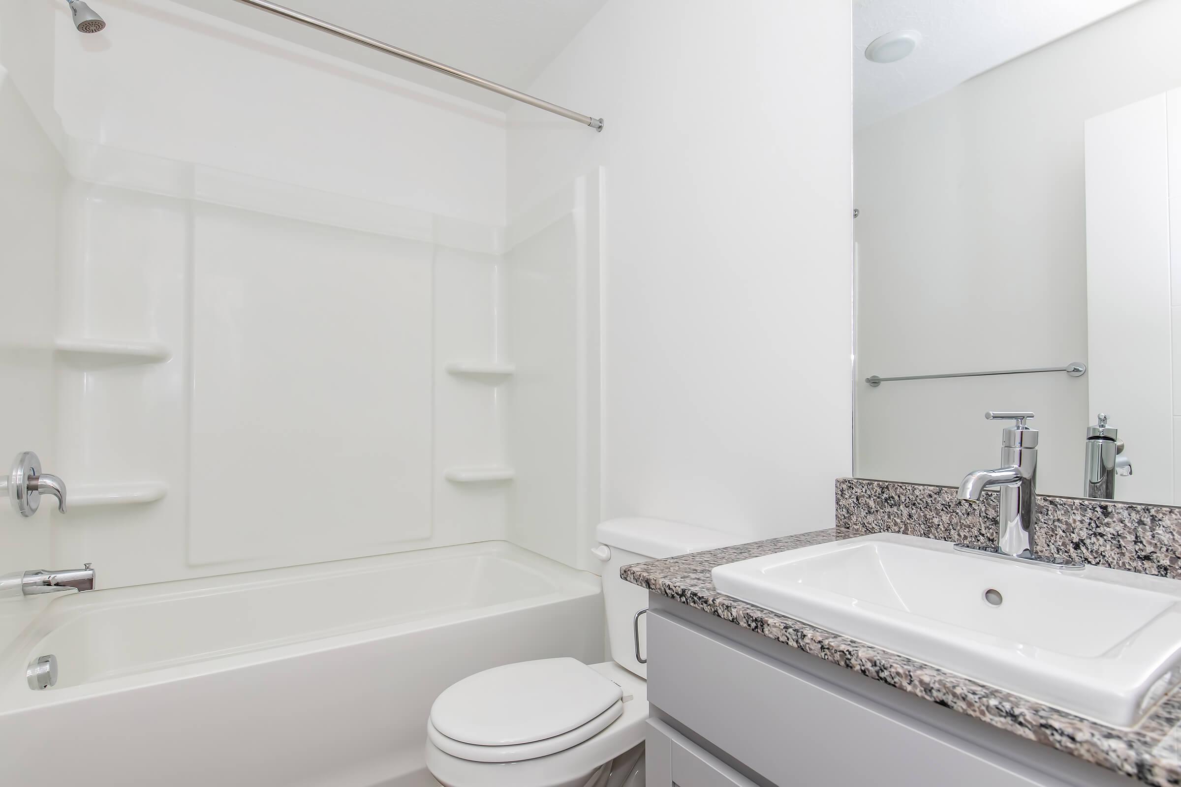 A clean and modern bathroom featuring a bathtub with a shower, a white sink with a sleek faucet, and a granite countertop. The walls are painted in light colors, and there is a large mirror above the sink, enhancing the bright and spacious feel of the room.