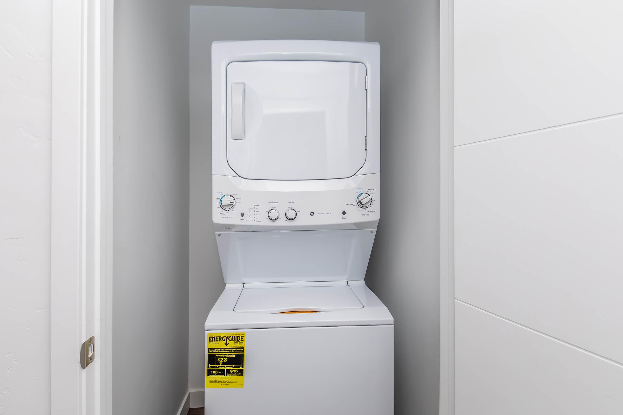 Stacked washer and dryer unit in a narrow laundry space, featuring control knobs and a small front-loading washer beneath a dryer. The walls are painted white, and there is a door on the left side leading out of the laundry area. A safety sticker is visible on the front.