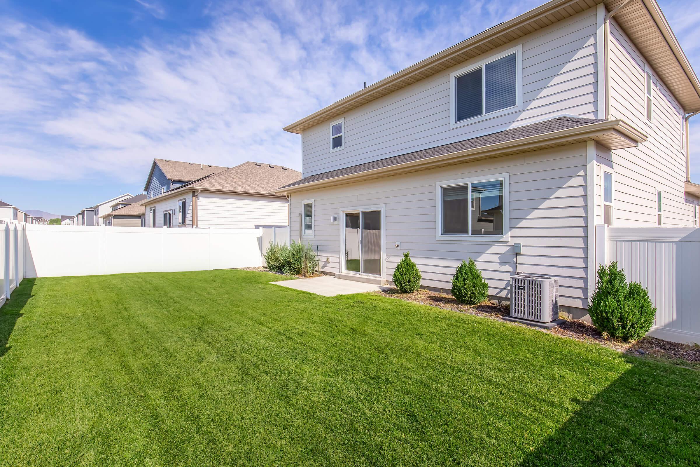 A well-maintained backyard featuring lush green grass, a concrete patio area, and small shrubs. The backdrop includes a two-story house with light-colored siding and several windows, bordered by a white fence. Clear blue skies with a few clouds complete the tranquil suburban setting.