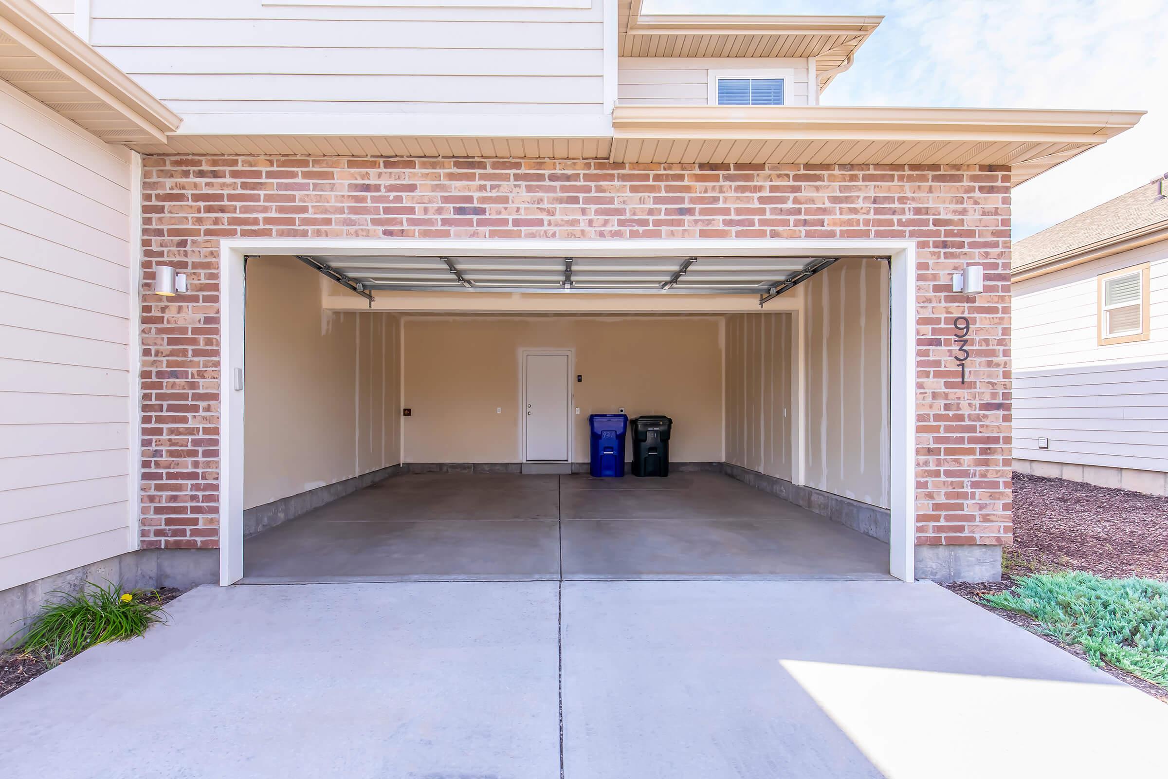 A clean, empty garage with a concrete floor and beige brick walls. Inside, there are two recycling bins, one blue and one black, positioned against the far wall. The garage door is open, revealing a sunny day outside. The house number 931 is displayed on the wall.