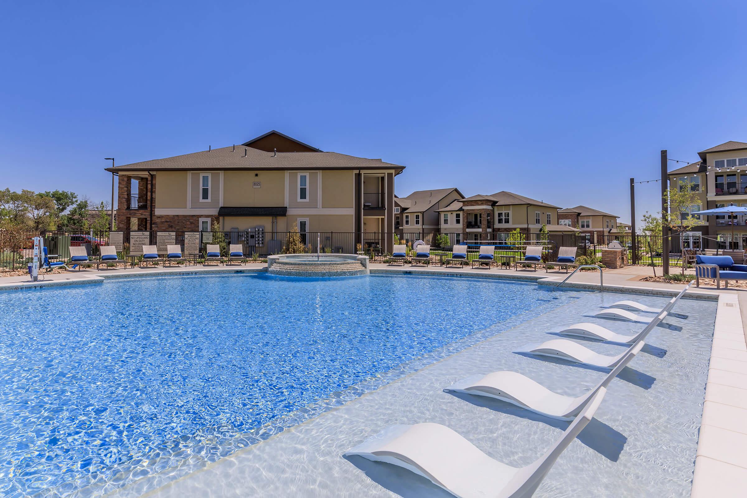 A bright, sunny day at an apartment complex featuring a sparkling blue swimming pool. Comfortable lounge chairs line the pool's edge, and modern buildings are visible in the background, surrounded by lush landscaping. The scene conveys a relaxing and inviting outdoor atmosphere.