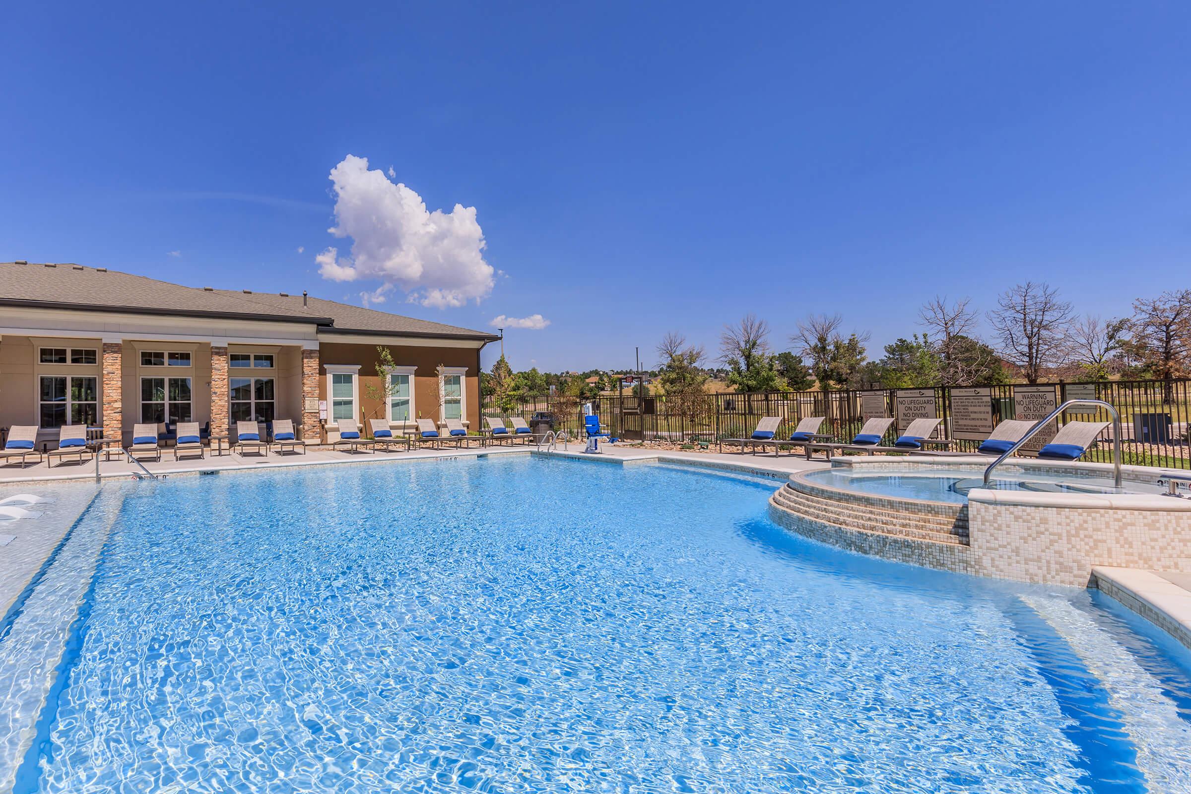 A bright blue swimming pool surrounded by lounge chairs under a clear sky. In the background, a well-maintained building with large windows and grassy areas is visible. A person is walking near the pool's edge. The scene conveys a relaxing outdoor atmosphere.