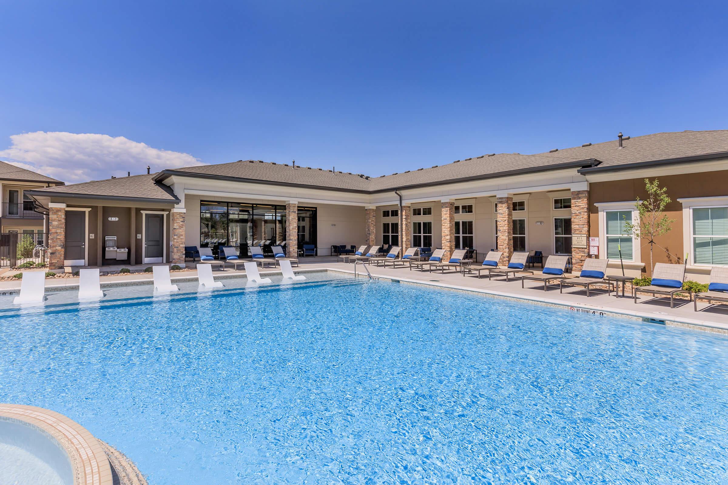 A clear blue swimming pool surrounded by lounge chairs and umbrellas, with a modern building in the background. The sky is sunny with a few clouds, and there are landscaped areas near the pool, creating a relaxing outdoor atmosphere.