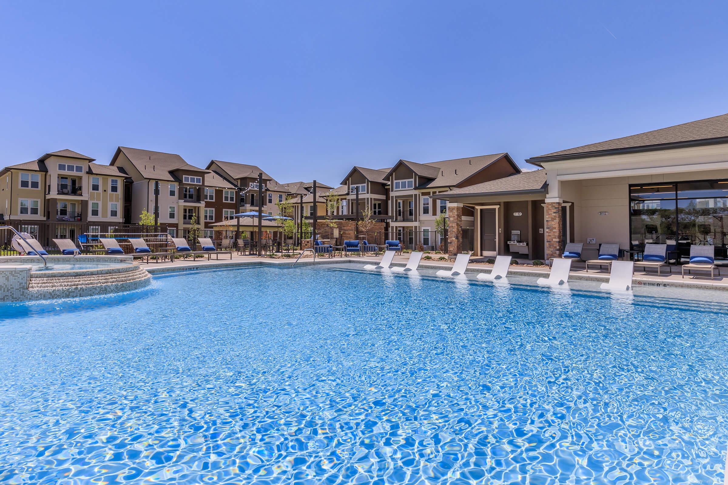 A sunny outdoor pool area with clear blue water, surrounded by lounge chairs and umbrellas. In the background, there are several multi-story residential buildings. The scene conveys a relaxed, upscale atmosphere ideal for leisure and socializing.