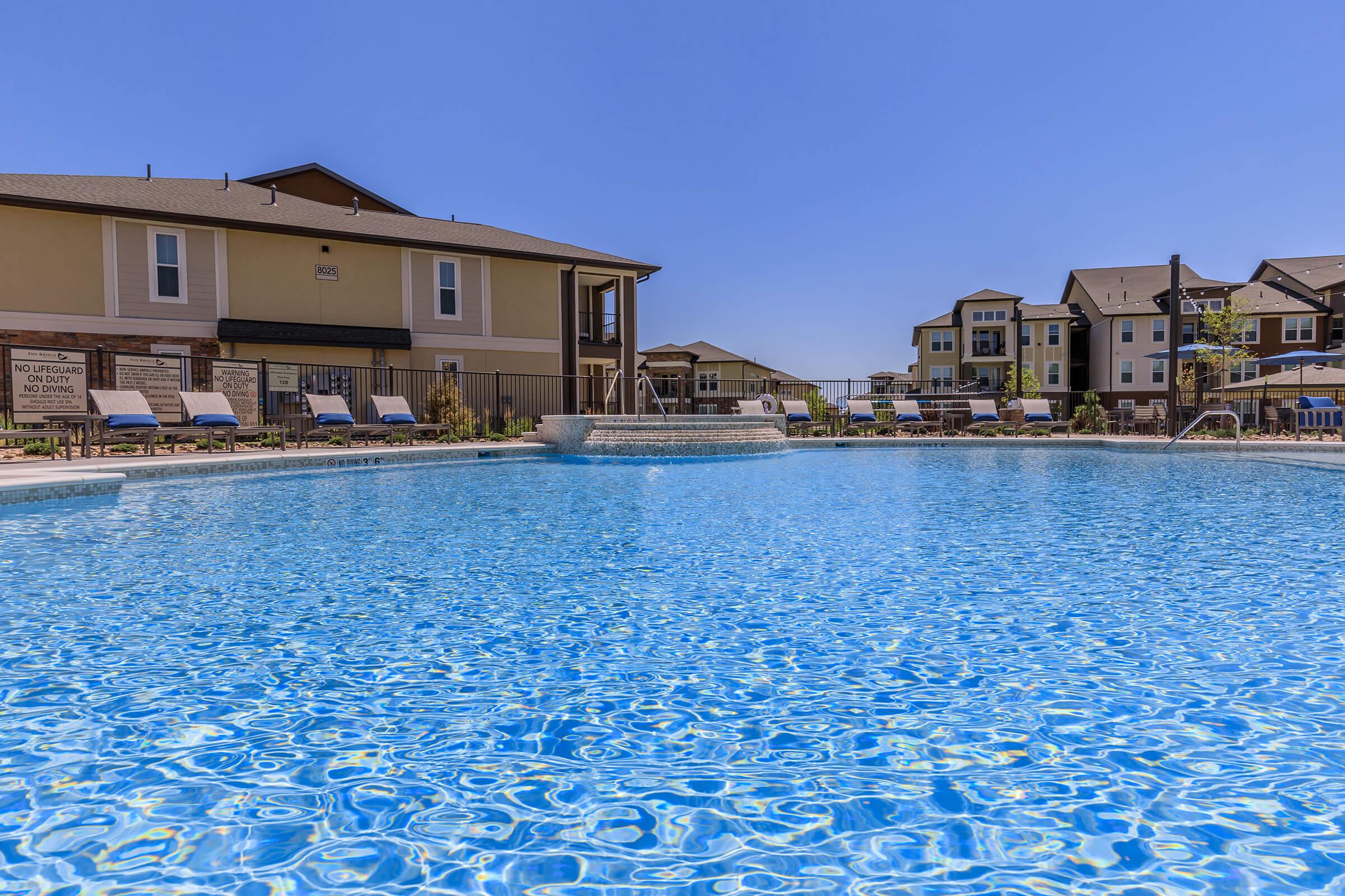 A clear blue swimming pool with lounge chairs around it, surrounded by modern residential buildings. The sky is bright and sunny, creating a vibrant atmosphere. The pool features a shallow area and a decorative water feature in the background.