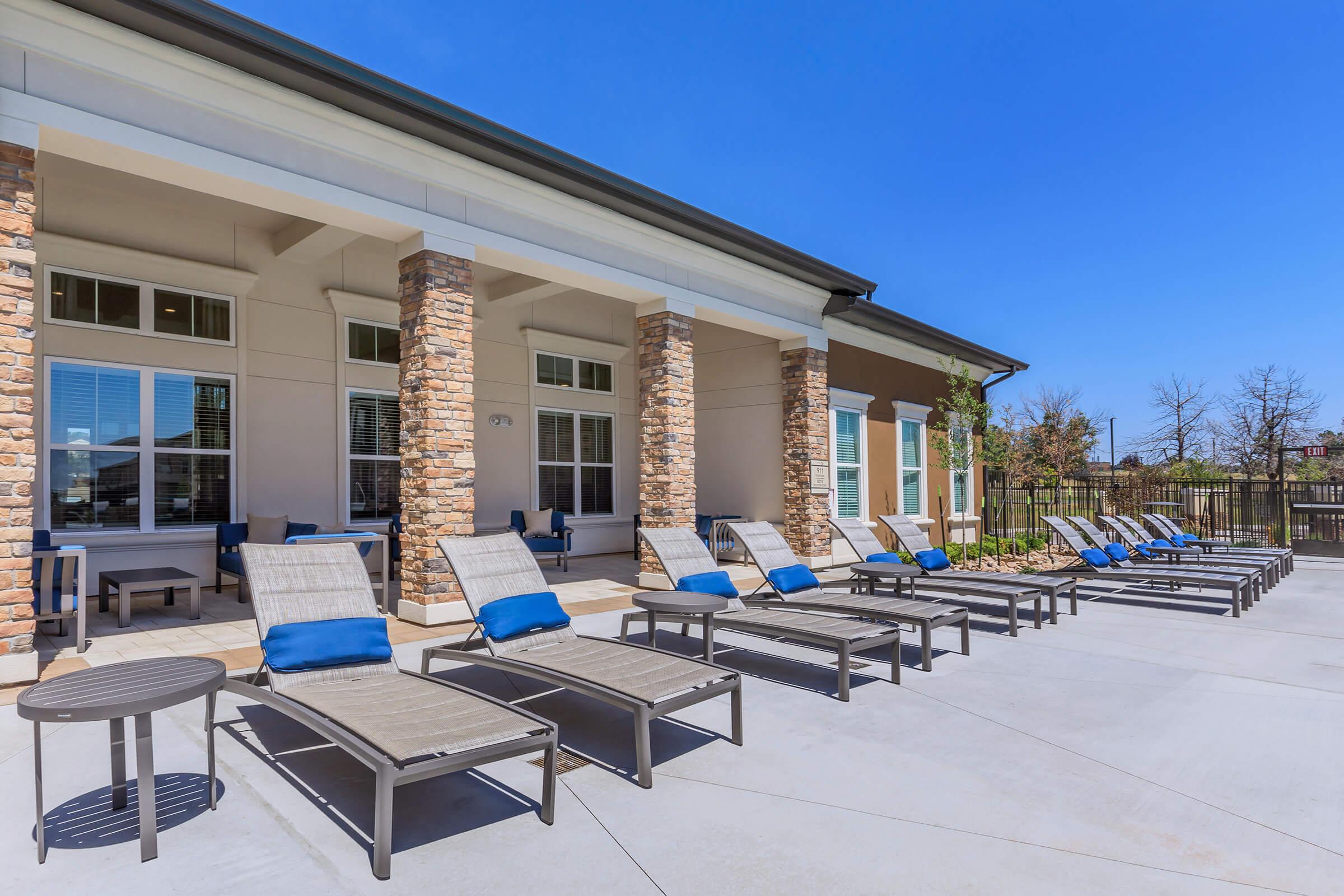 A sunny outdoor area featuring several reclining lounge chairs with blue cushions, arranged neatly by a modern building. The space includes a stone facade, large windows, and a clear blue sky, creating a relaxing atmosphere.