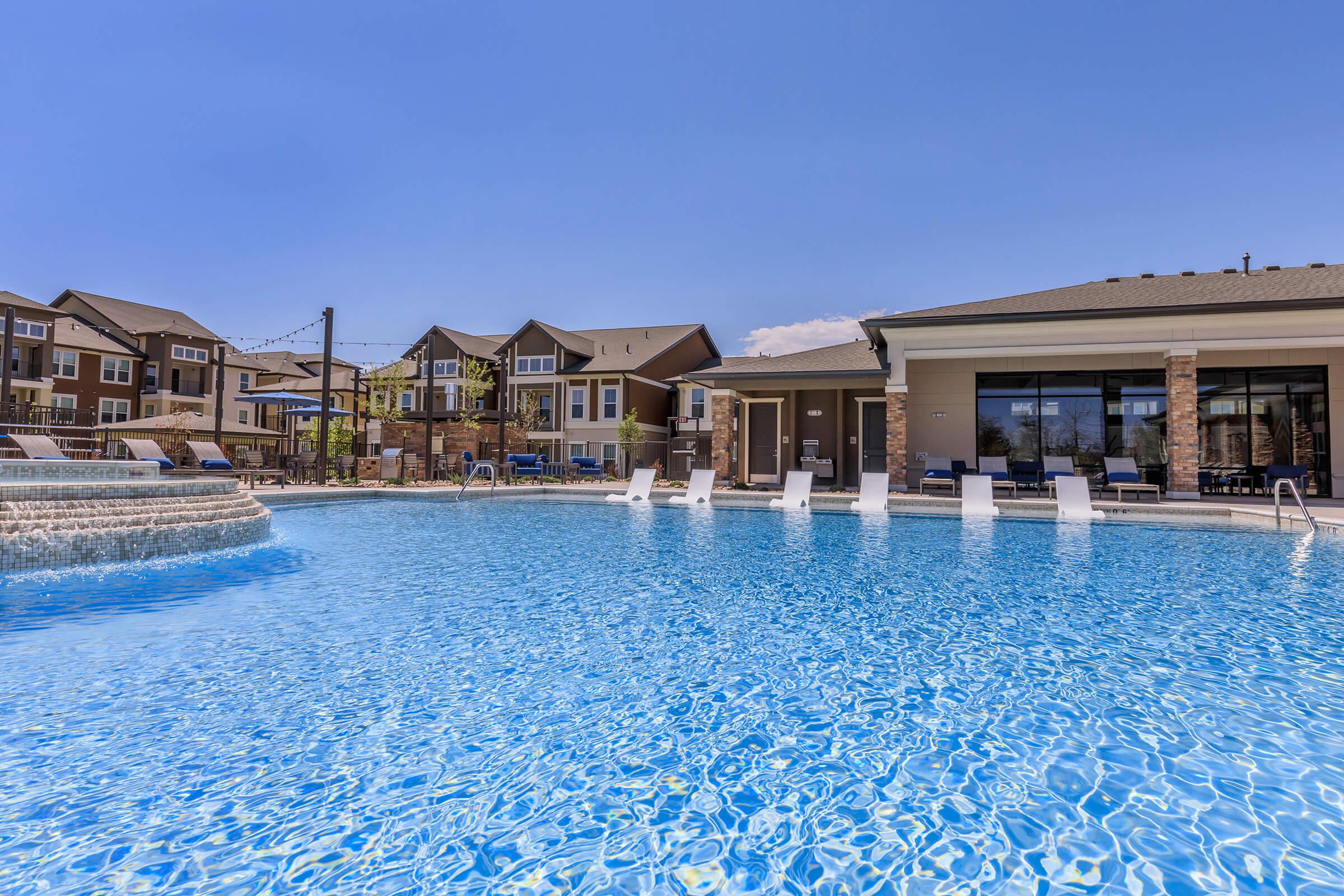 A sparkling blue pool with a cascading fountain, surrounded by lounge chairs and a sunlit patio. In the background, modern apartment buildings with various colors can be seen under a clear blue sky. The scene conveys a relaxing outdoor space perfect for leisure and recreation.