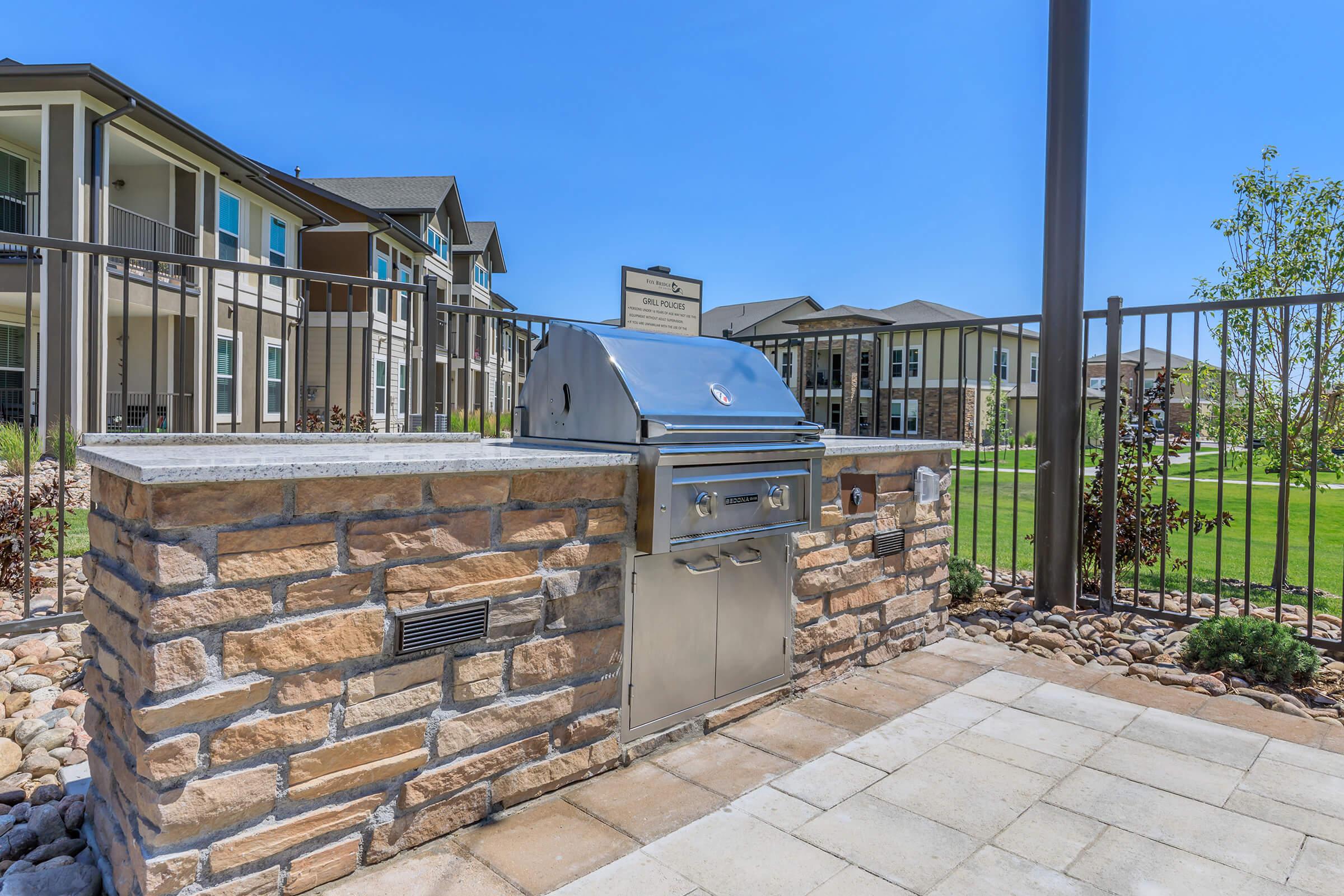 Outdoor barbecue grill featuring a stainless steel design, positioned on a stone base with a paved area, surrounded by green grass and residential buildings in the background. A tall fence encircles the area, providing a private outdoor cooking space. Clear blue sky above enhances the setting.