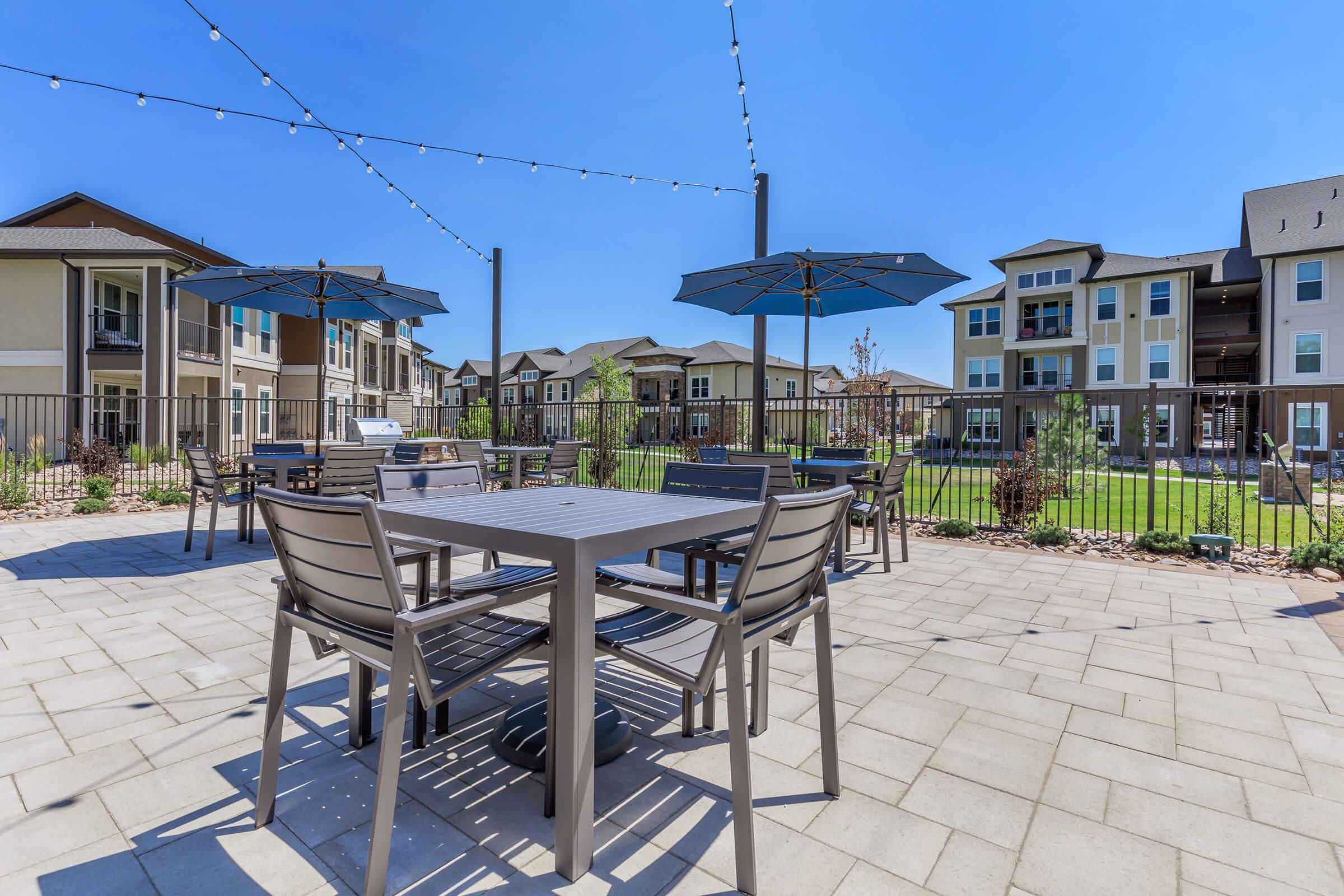 Outdoor area featuring a patio with tables and chairs, surrounded by green grass and buildings. The scene includes large umbrellas providing shade, and decorative string lights overhead in a bright blue sky. Ideal for relaxation and gatherings in a residential setting.