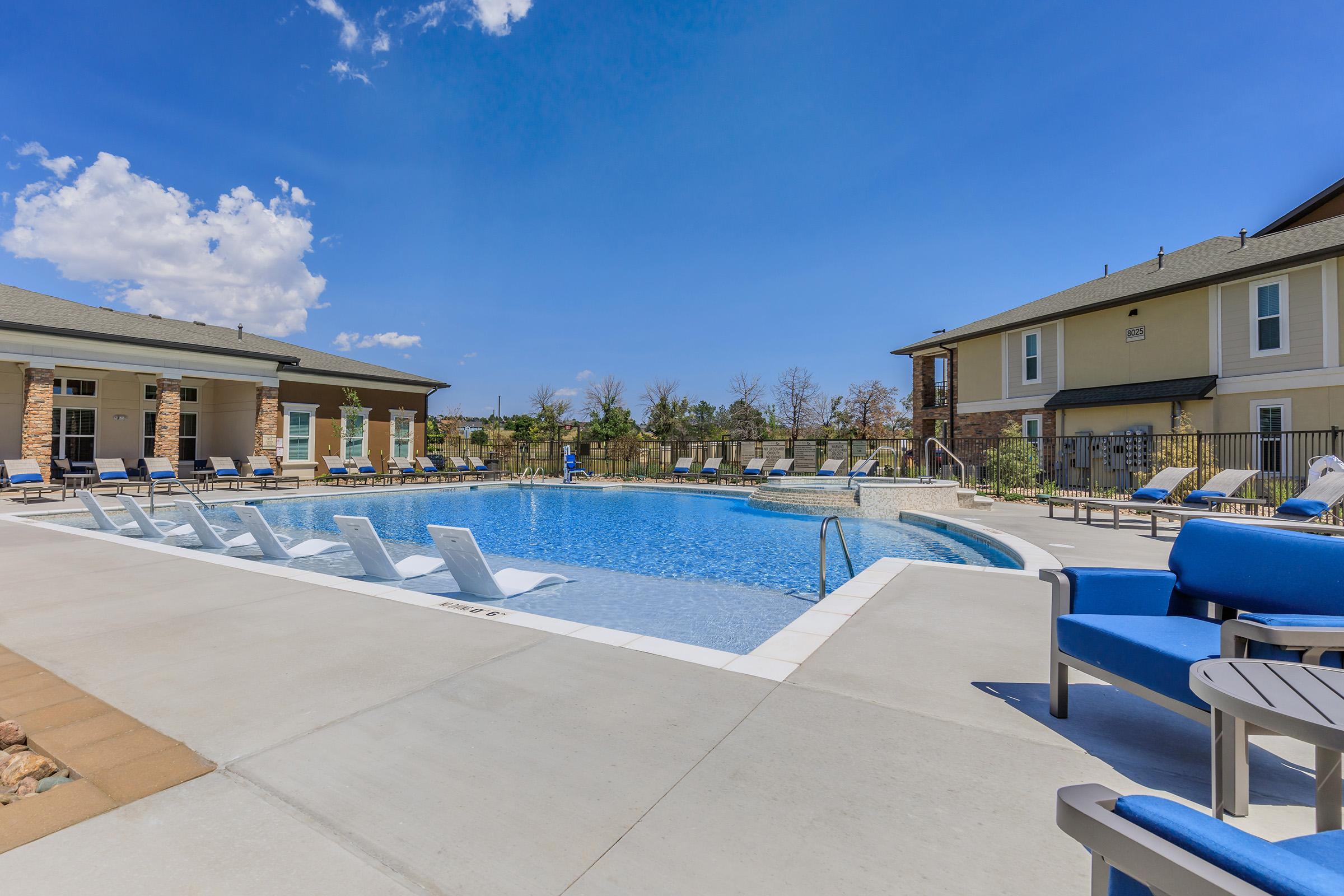 A tranquil outdoor swimming pool surrounded by lounge chairs, with a clear blue sky and a few fluffy clouds overhead. Nearby buildings are visible, suggesting a residential or leisure area. The scene conveys a inviting atmosphere for relaxation and enjoyment.
