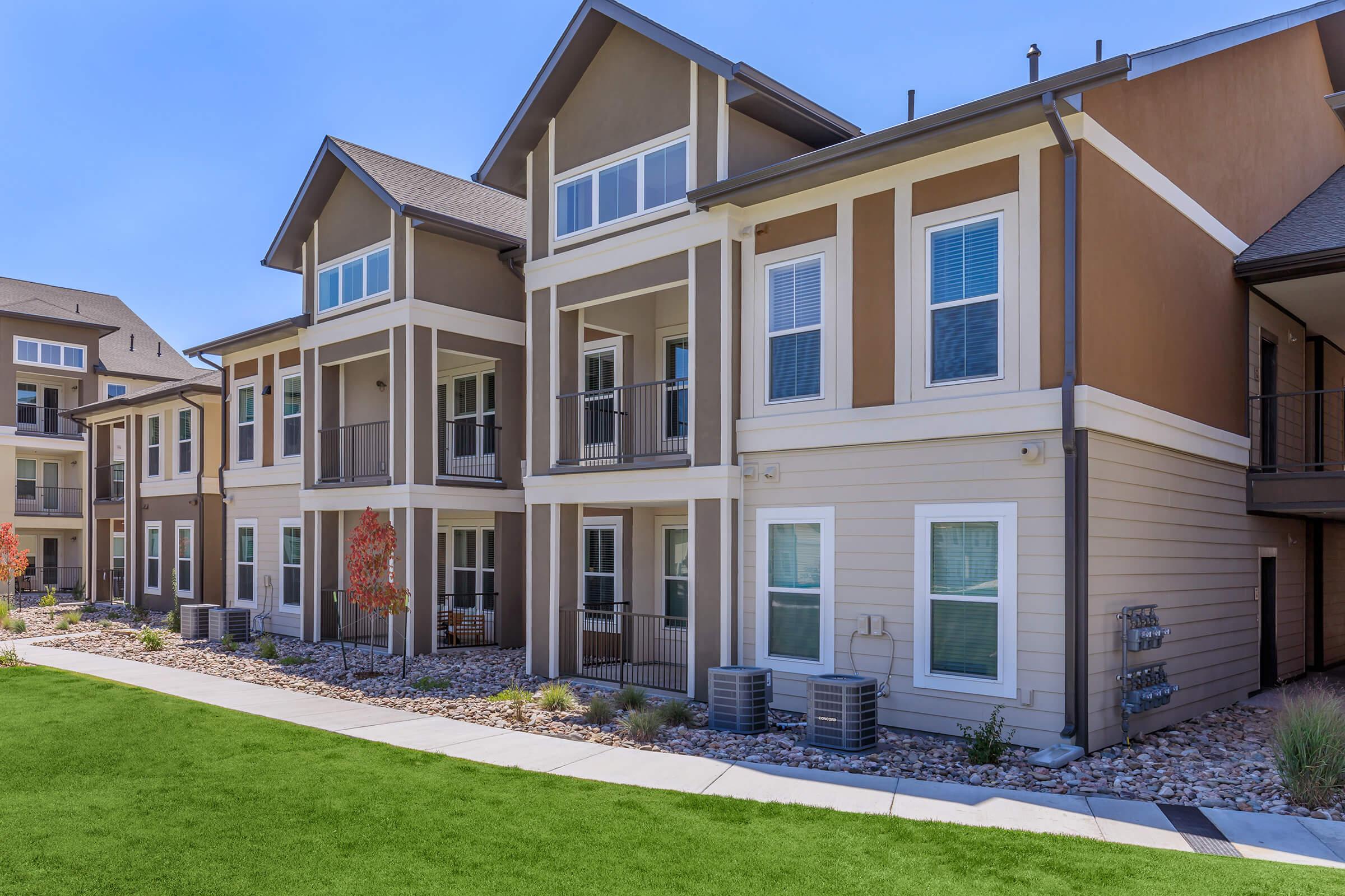 Modern apartment complex with multiple stories featuring balconies and large windows. The exterior is a mix of brown and beige siding, surrounded by green lawns and landscaped areas with small trees. There are air conditioning units visible near the ground level. Clear blue sky overhead.