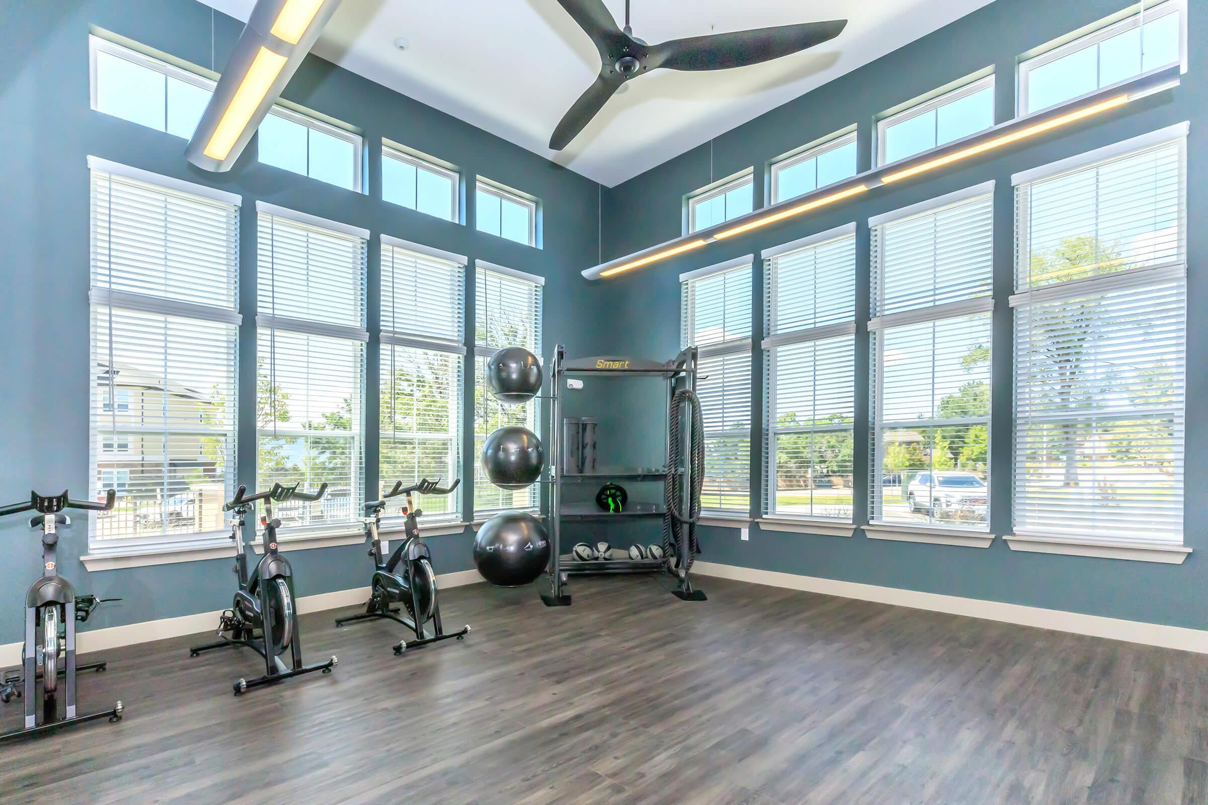 A bright, modern fitness room featuring several exercise bikes, a cable machine, and fitness balls. Large windows allow natural light, and a ceiling fan provides ventilation. The walls are painted a calming blue, enhancing the tranquil atmosphere for workouts.