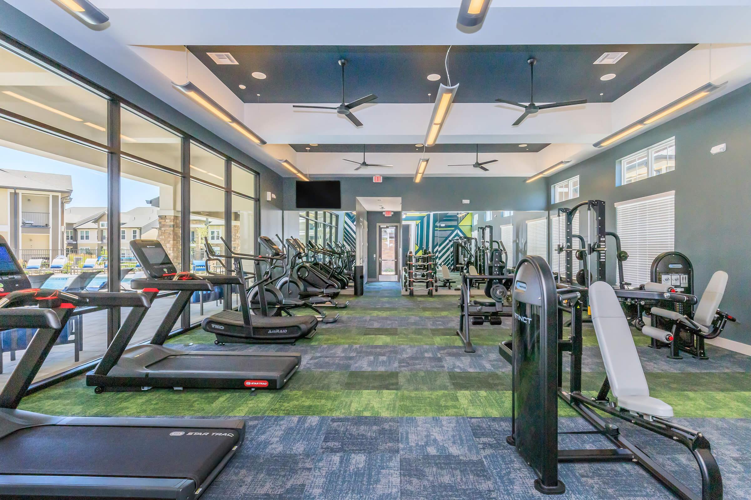 A modern gym interior featuring various exercise equipment, including treadmills, elliptical machines, and weight machines. Large windows allow natural light to fill the space, and the flooring is a mix of carpet and rubber. The design is contemporary with ceiling fans for ventilation.