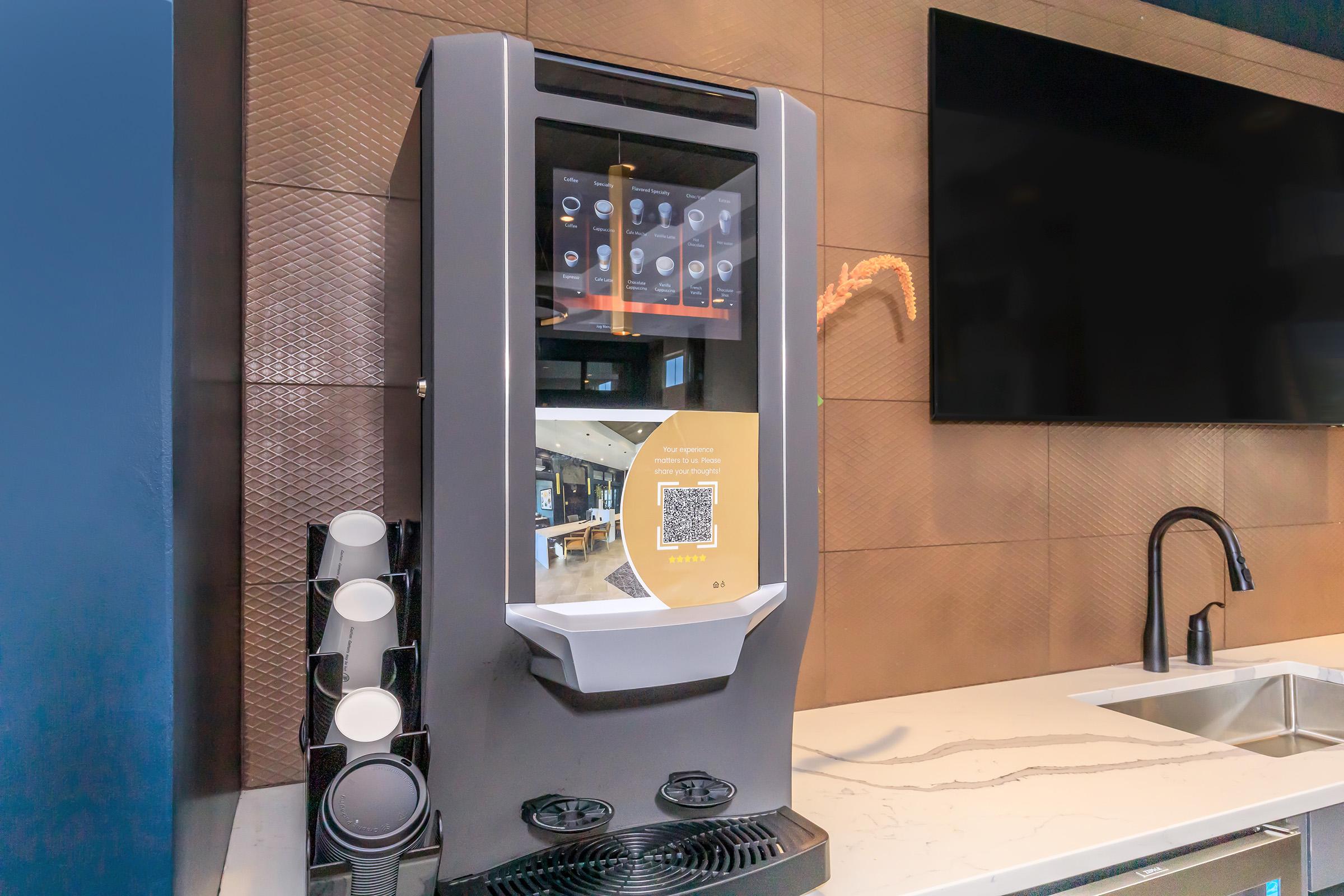 A modern beverage dispenser featuring a digital display, located in a sleek kitchen environment. The dispenser is mounted on a wall with a stone texture, and includes a QR code for usage instructions, alongside several beverage options visible through the glass front. A sink is also present nearby.