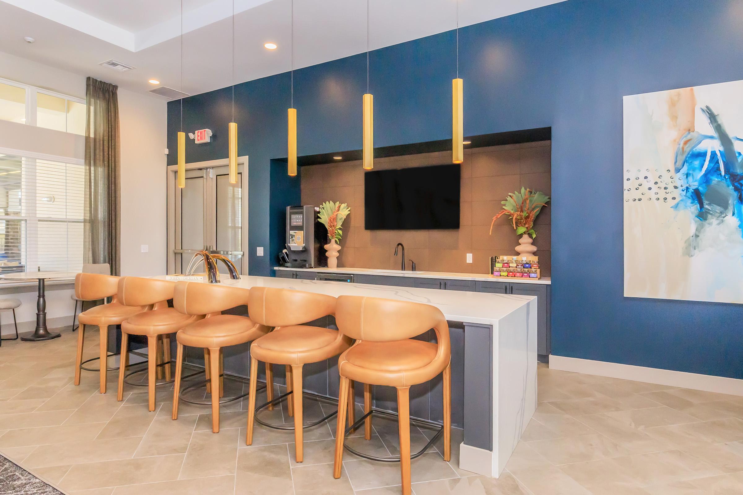 Bright and modern kitchen bar area with tan leather stools, a dark blue accent wall, and decorative plants. Features include a large countertop, a wall-mounted television, and a stylish piece of artwork. Sunlight streams in from large windows, enhancing the inviting atmosphere.