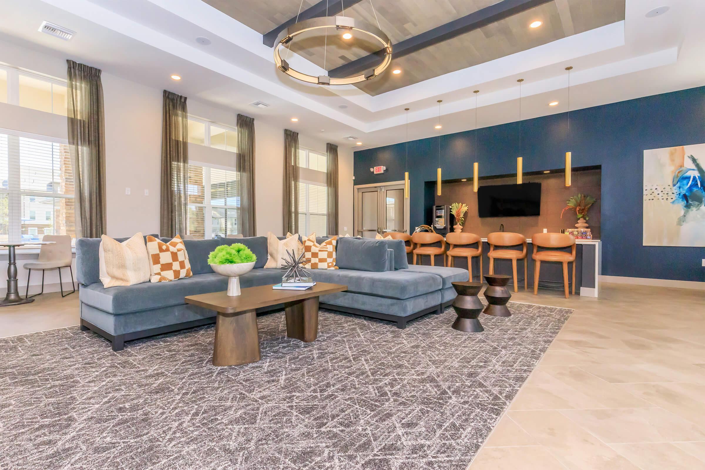 A spacious and modern lounge area featuring a gray sectional sofa with decorative pillows, a coffee table, and two accent stools. The room has large windows allowing natural light, a stylish light fixture, and a dark accent wall with a mounted TV. The decor includes plants and artwork, creating a welcoming atmosphere.