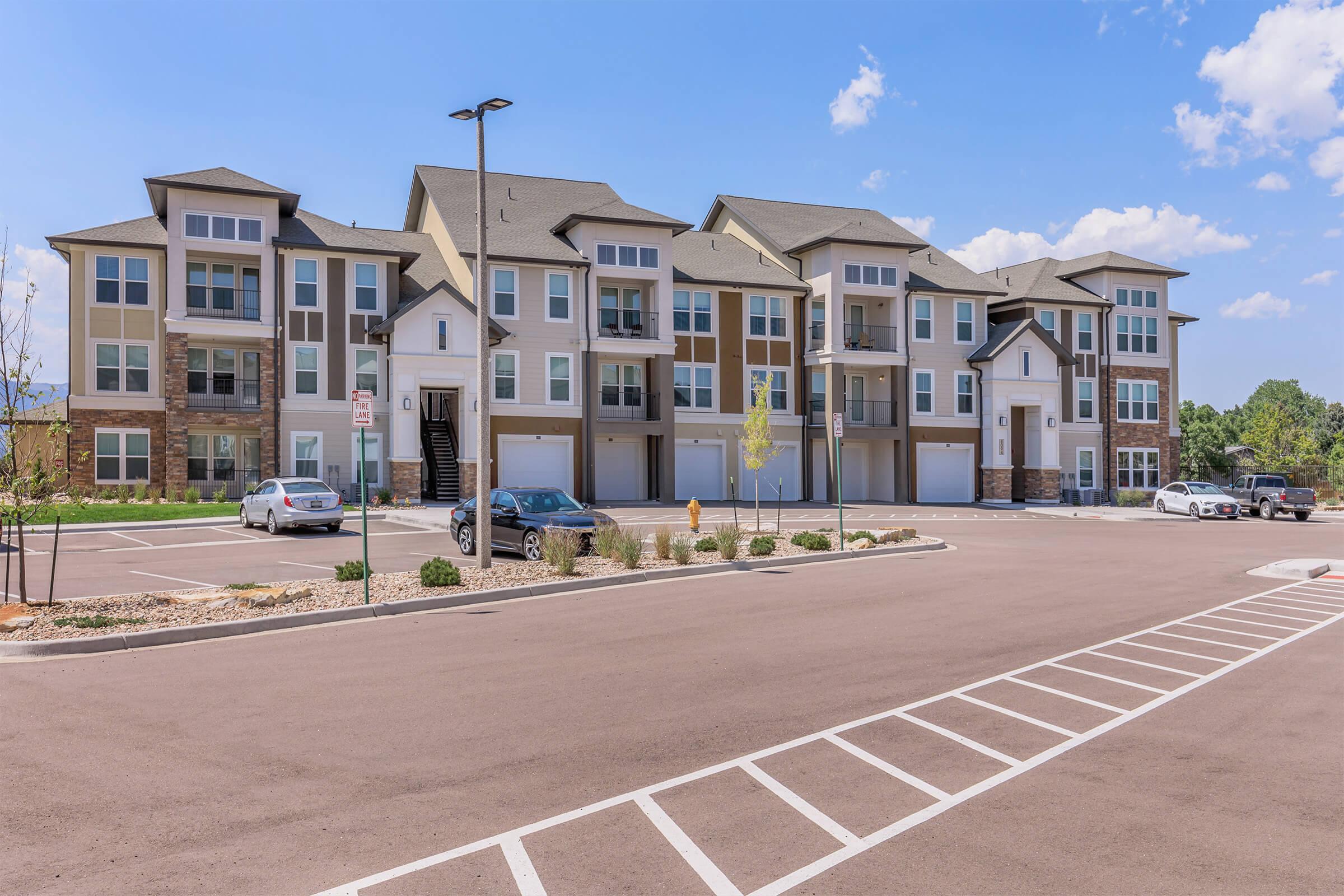 A modern apartment complex featuring multiple three-story buildings with a mix of stone and siding exteriors. The parking lot in front has several parked cars and is lined with landscaped areas, under a clear blue sky with a few clouds.