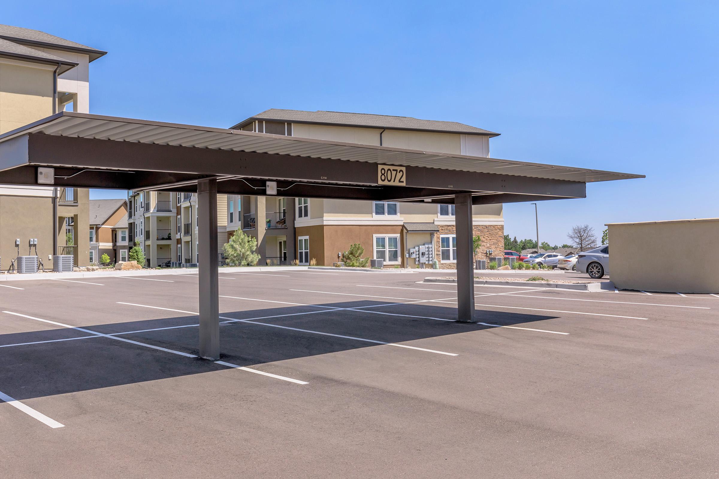 A parking lot with a covered carport structure. The carport features the number "8072" on it. Surrounding the area are modern apartment buildings, with clear blue skies overhead and a few parked cars visible in the background. The pavement is clean and recently paved.