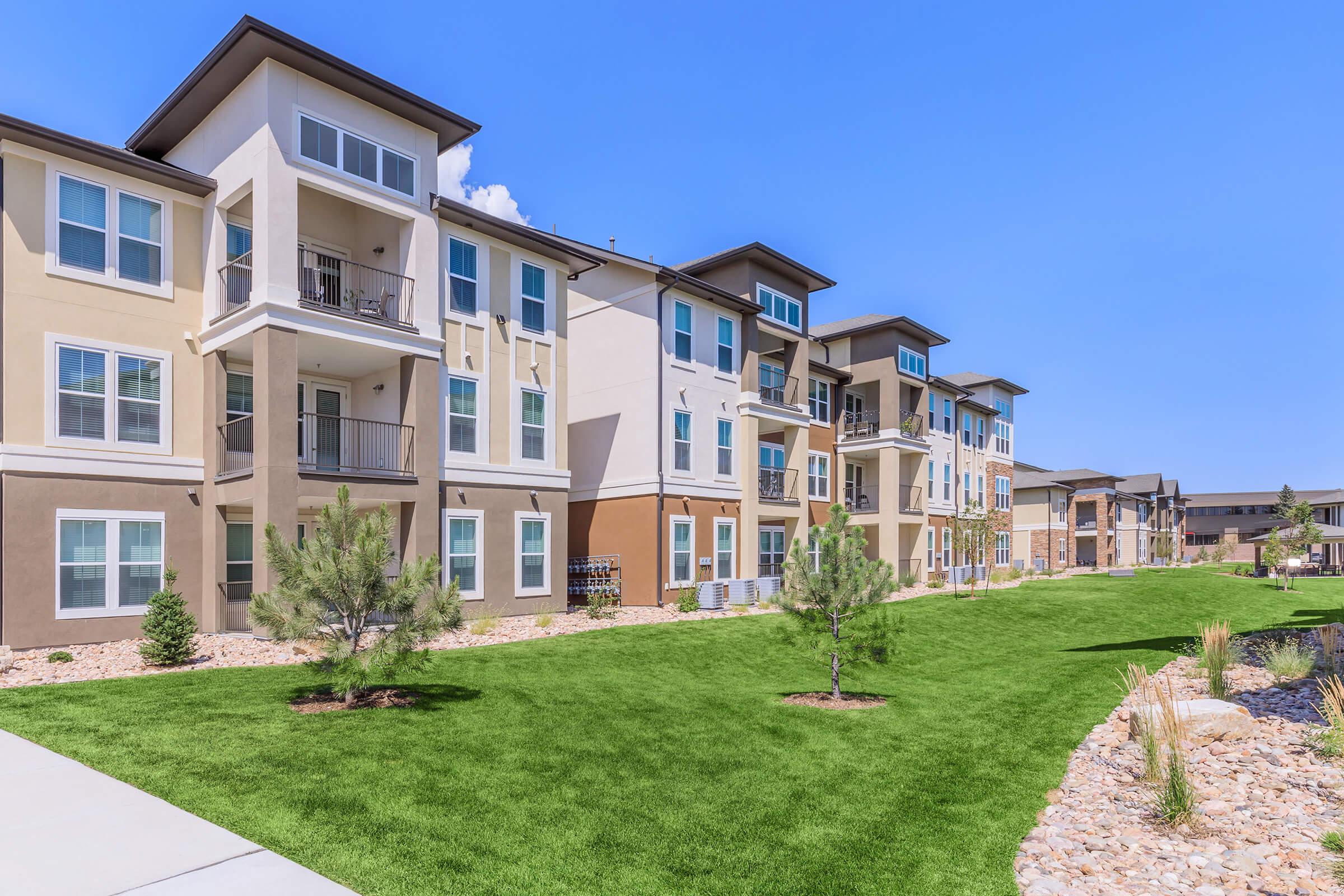 A row of modern, multi-story apartment buildings with balconies, surrounded by well-maintained green lawns and low shrubs under a clear blue sky.