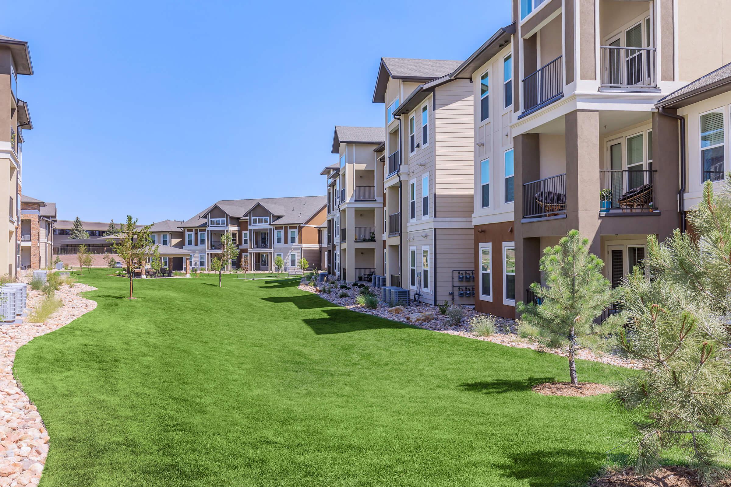 A well-maintained apartment complex featuring modern buildings with balconies, surrounded by lush green lawns and landscaped areas. Clear blue skies enhance the inviting atmosphere, making it a pleasant outdoor space for residents.