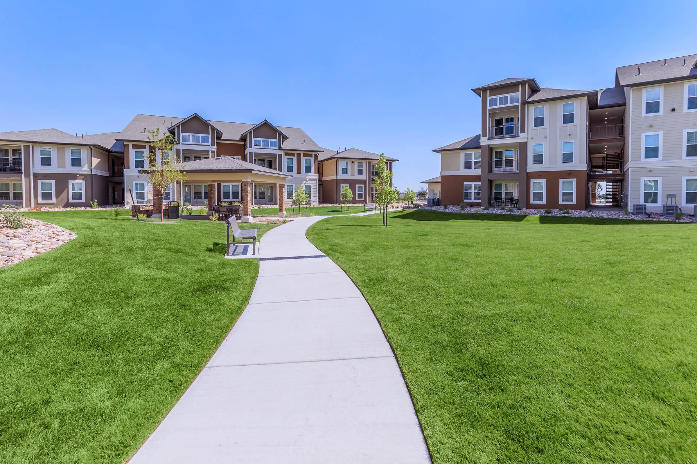 A well-maintained residential complex featuring several apartment buildings with modern architecture. A paved pathway winds through lush green lawns, leading to a gazebo. Clear blue skies create a bright atmosphere, enhancing the inviting outdoor space for residents to enjoy.