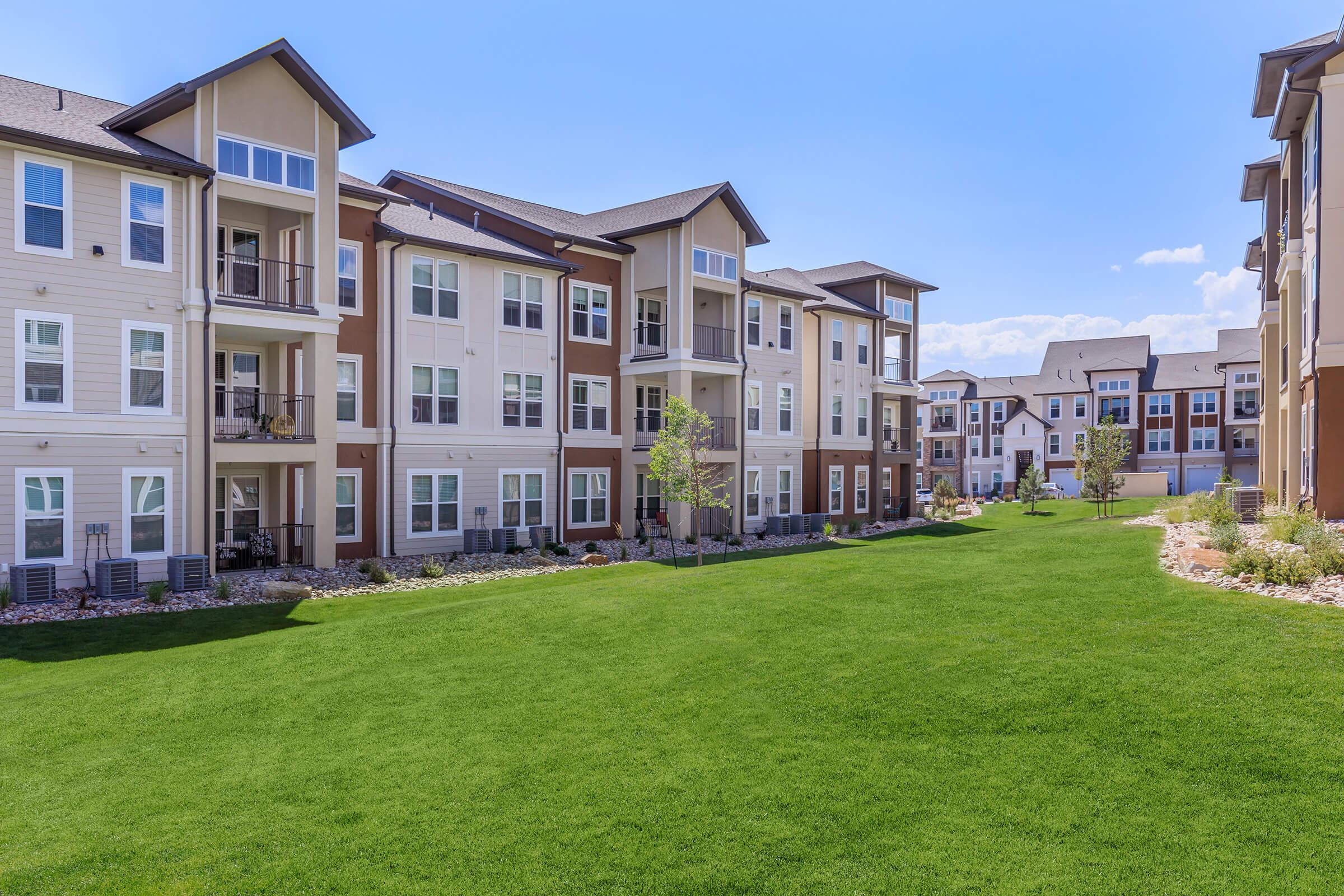 A well-maintained apartment complex featuring multiple three-story buildings with a modern design. The buildings have balconies and large windows, surrounded by a lush green lawn and landscaping. Clear blue skies provide a bright atmosphere, emphasizing the inviting outdoor space.