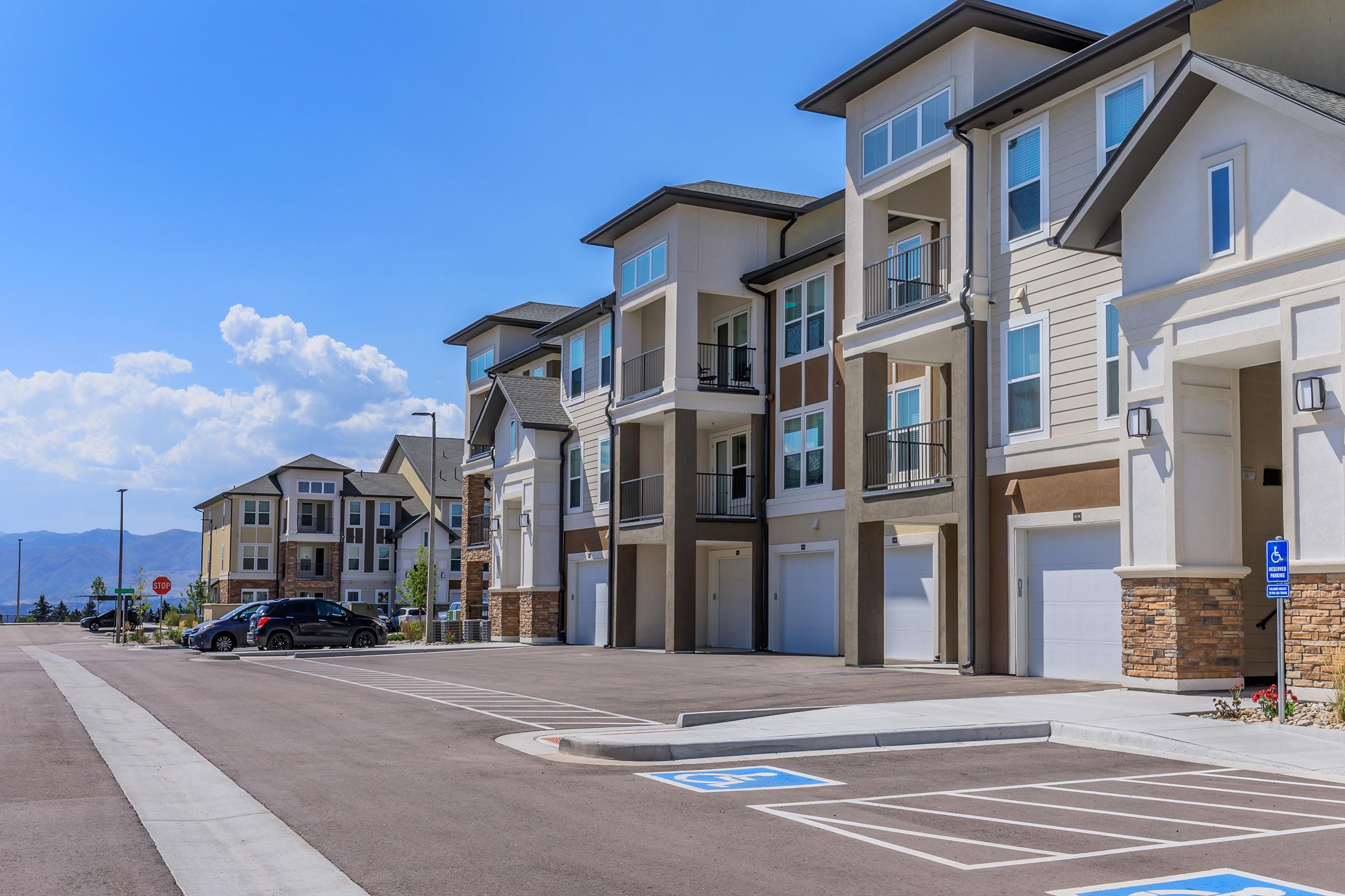 Residential buildings lined along a well-paved street with parking spaces, featuring modern architecture and balconies. Clear blue sky with a few clouds in the background, showcasing a serene neighborhood setting.