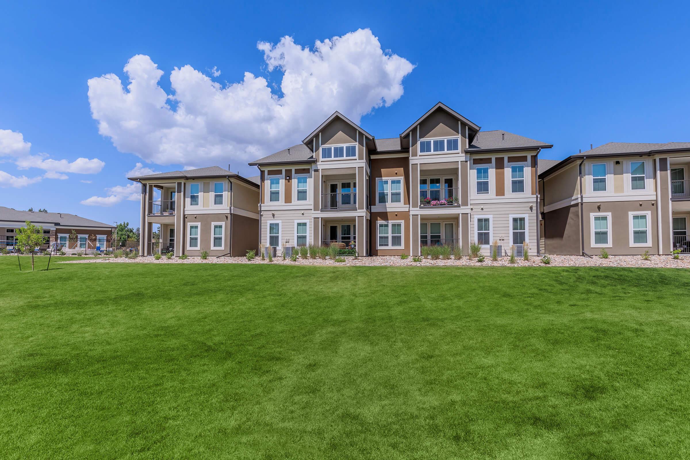 A multi-unit residential building set against a bright blue sky with fluffy white clouds. The lawn in front is lush and green, featuring well-maintained landscaping. The building showcases a modern architectural style with balconies and large windows.