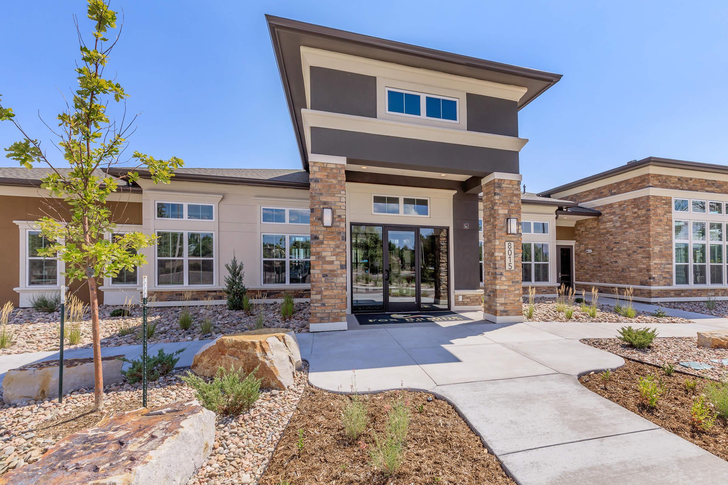 Modern building entrance with a stone and stucco facade. The pathway leads to a large glass door, flanked by windows. Surrounding the entrance are landscaped areas with rocks and small plants, creating a welcoming atmosphere. Bright blue sky enhances the overall appeal of the setting.