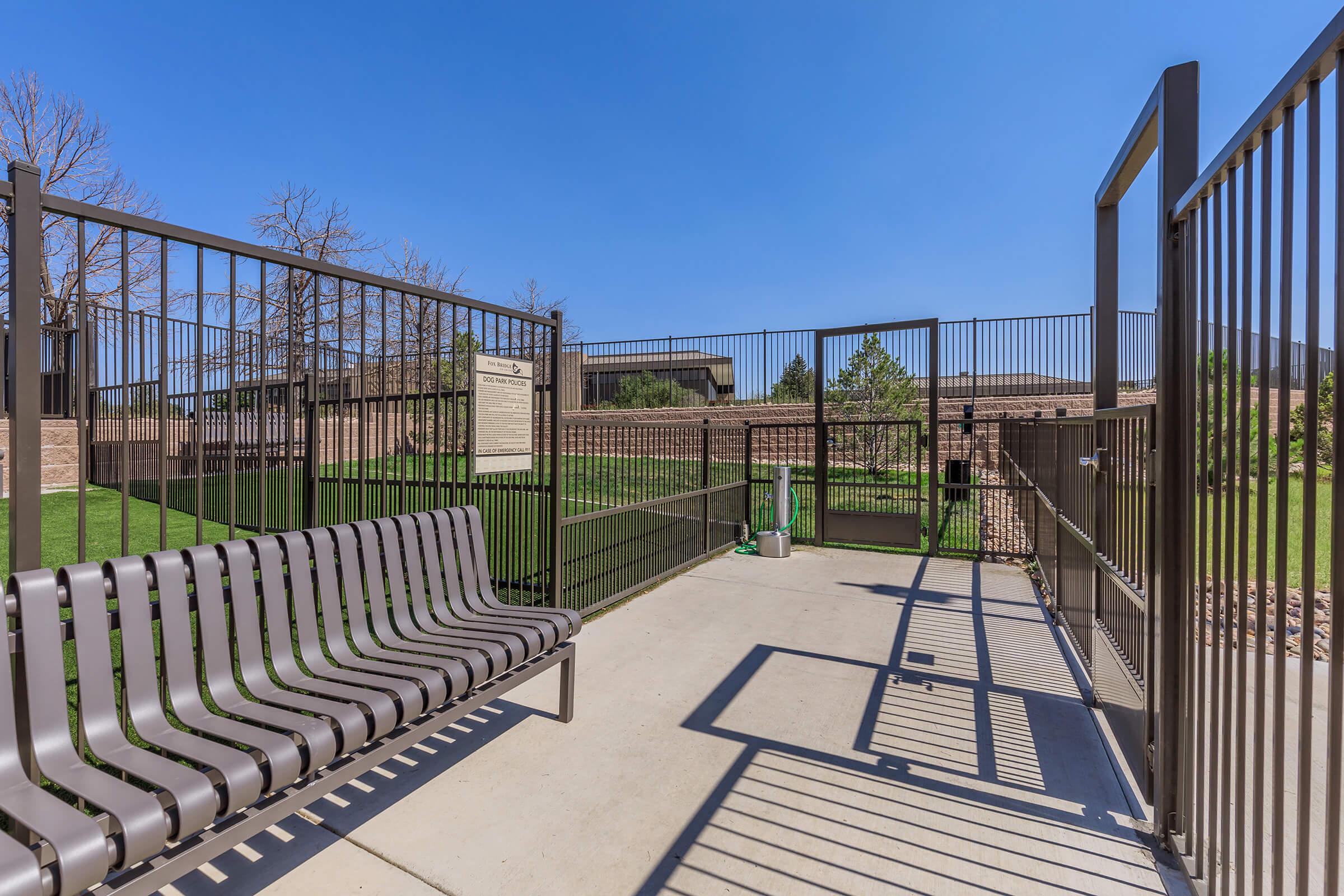 A fenced outdoor dog park area featuring a concrete surface, a bench for seating, and grassy patches in the background. The space is enclosed with metal fencing, and there's a gate visible. The sky is clear and blue, indicating a sunny day.