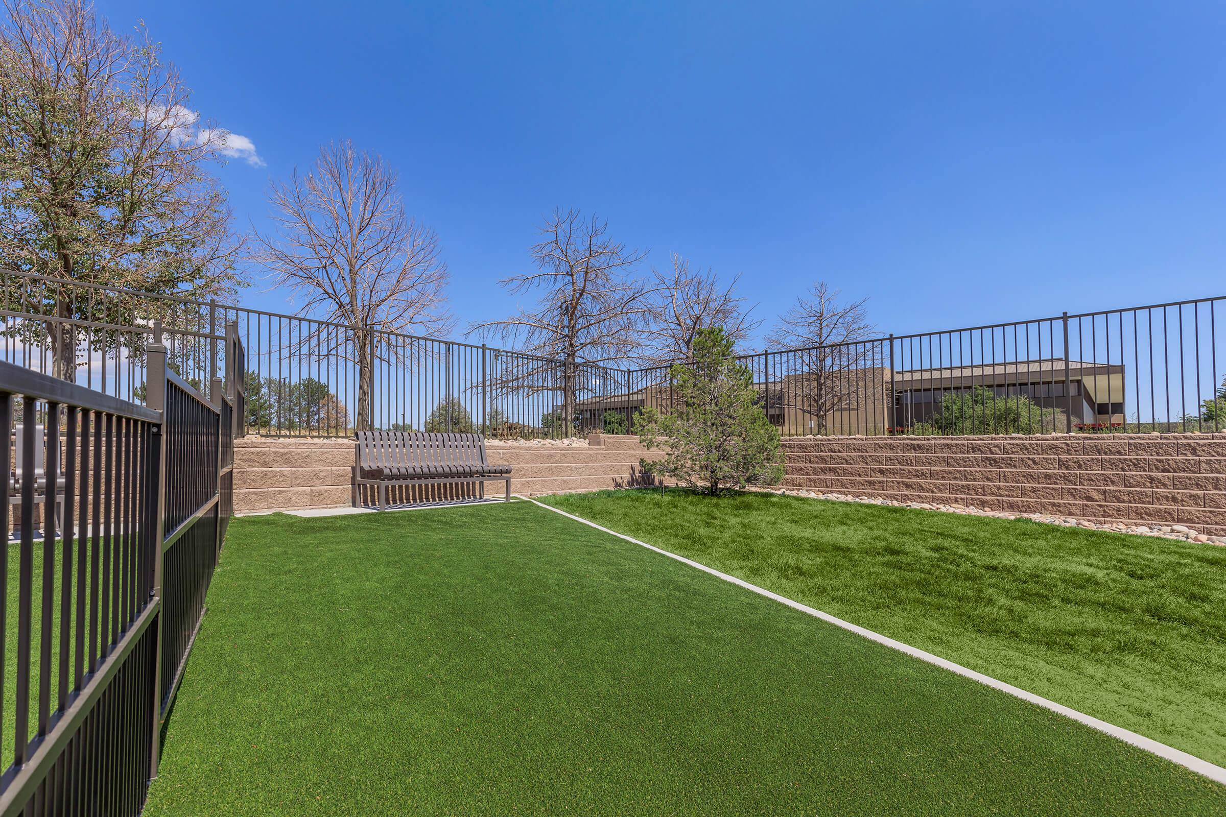 A landscaped outdoor area features artificial turf, surrounded by a black fence. In the background, there are sparse trees and a bench along the edge. The sky is clear and blue, creating a serene environment suitable for relaxation or recreation.