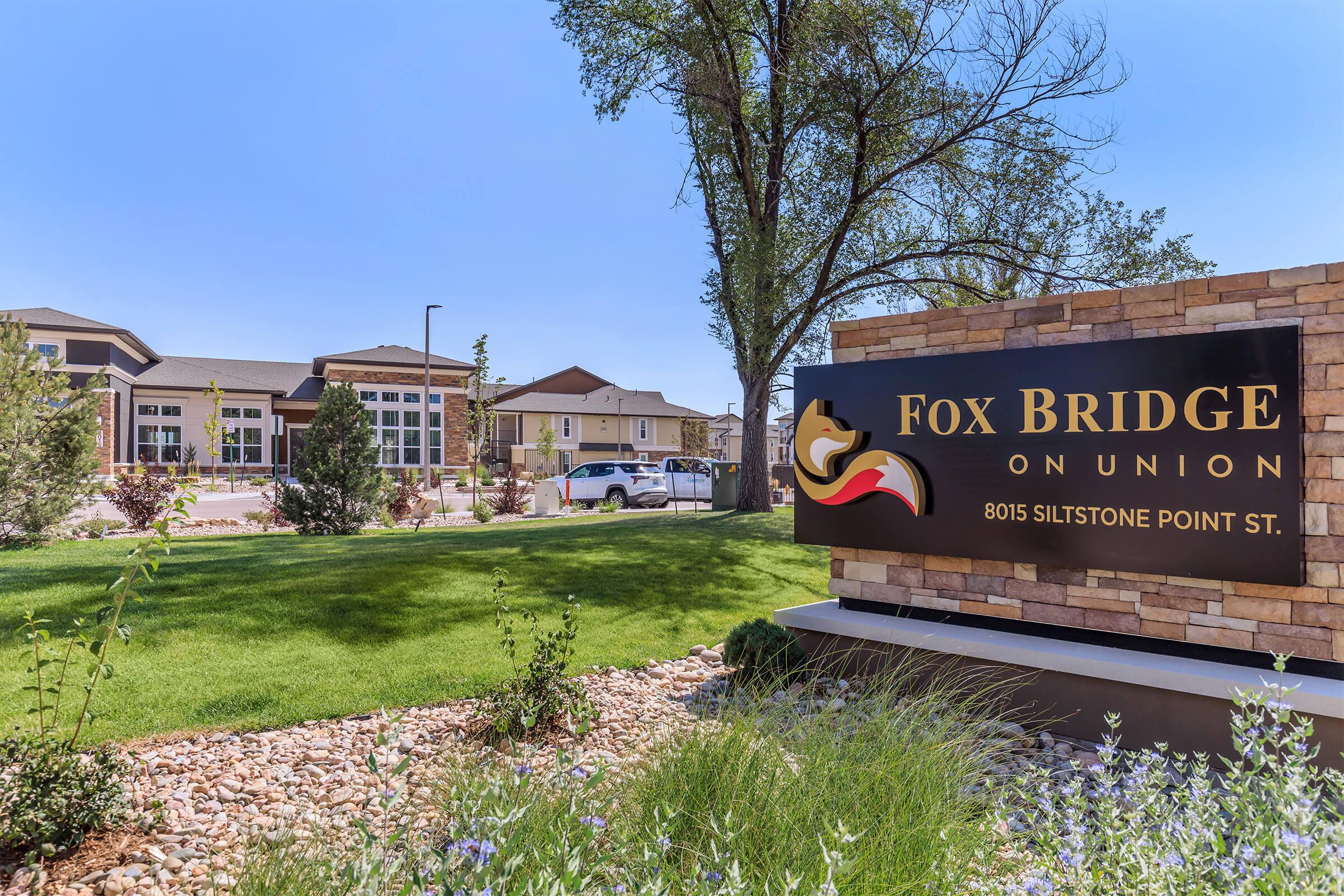Sign for Fox Bridge on Union, displaying the name prominently. The image also shows well-maintained landscaping with green grass and decorative rocks. In the background, there are modern residential buildings under a clear blue sky.