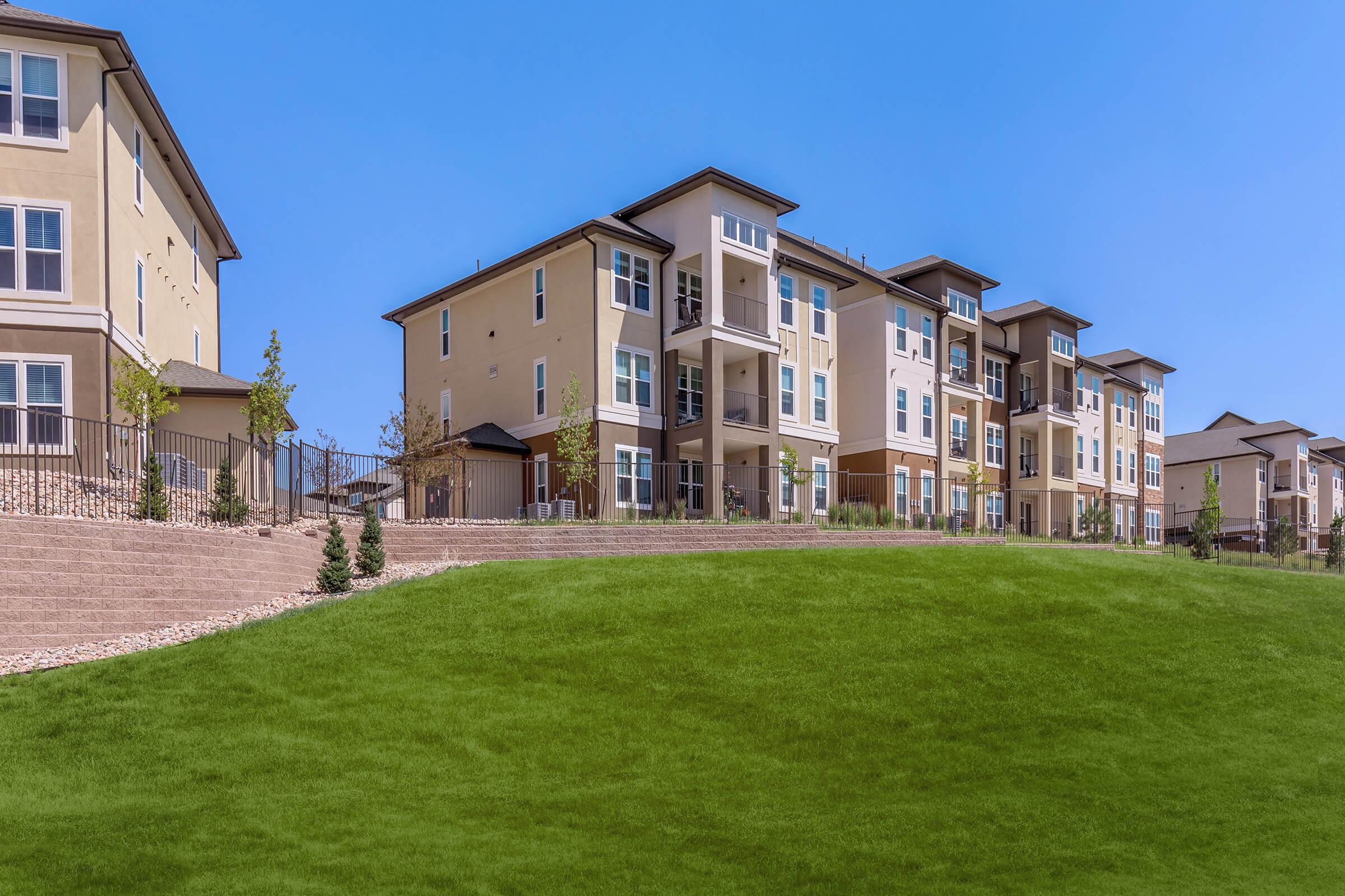 Row of modern multi-story apartment buildings on a gently sloping green hillside. The buildings feature balconies and a contemporary architectural design, with a clear blue sky in the background. Small landscaped areas and retaining walls enhance the outdoor space around the properties.