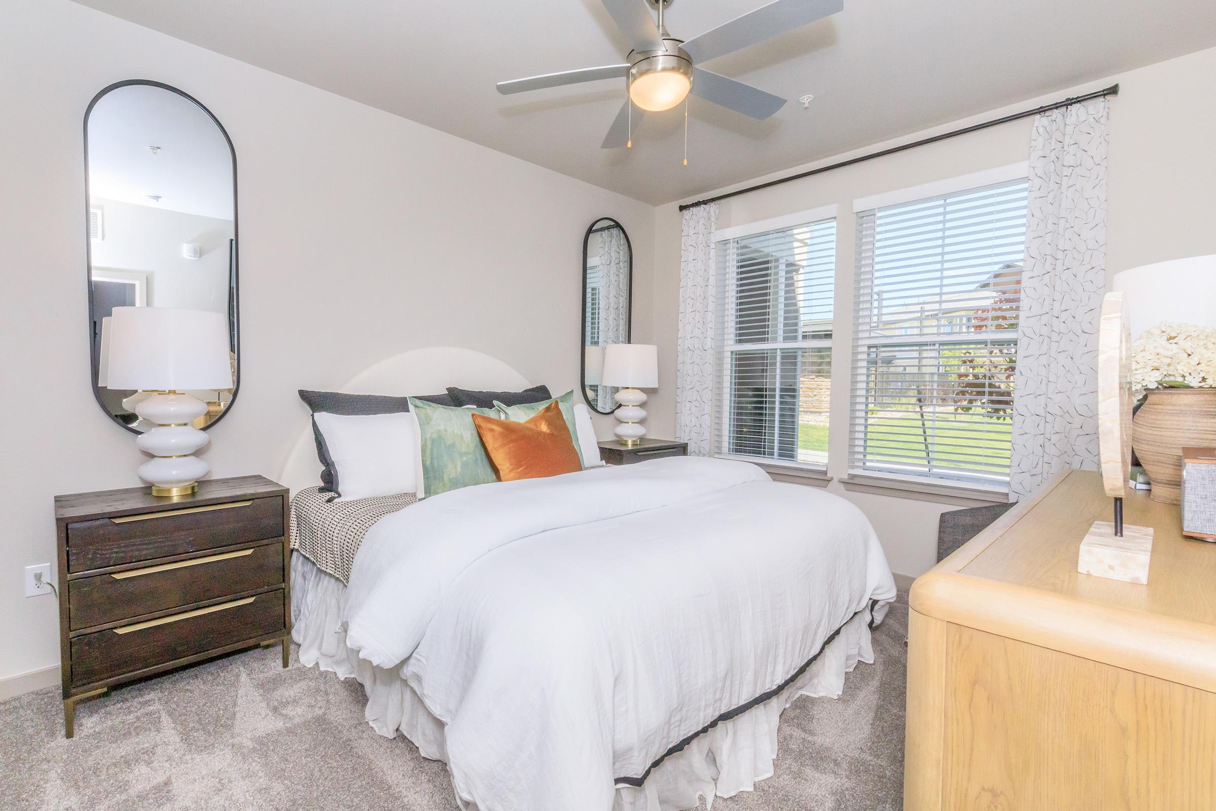 A cozy bedroom featuring a comfortable bed with white bedding and decorative pillows. It has two stylish mirrors on the walls, a ceiling fan, and bedside tables with modern lamps. Natural light streams in through a window overlooking a grassy area. The decor is neutral with touches of color for a warm ambiance.