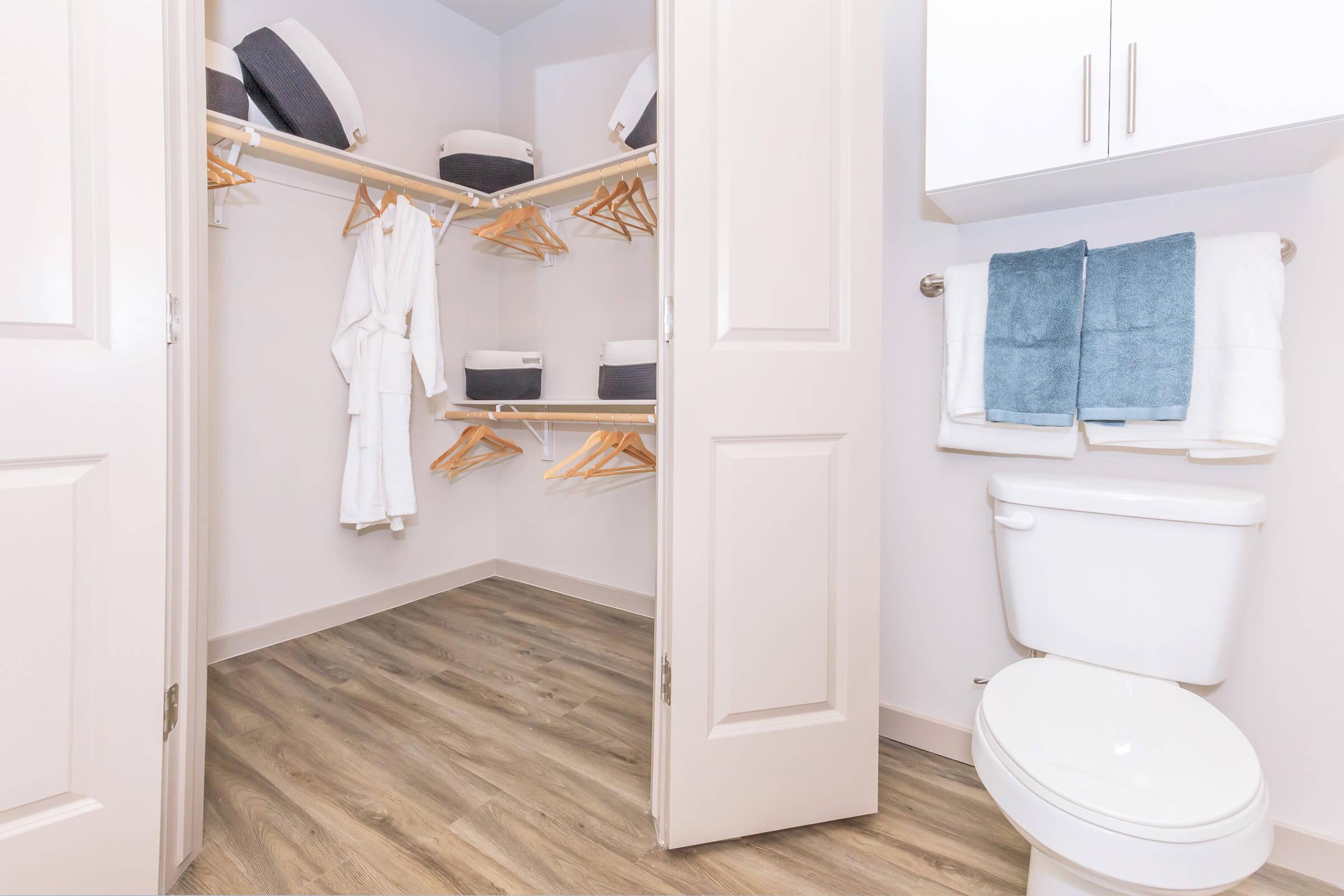 A tidy, organized bathroom closet with wooden shelving. The shelves hold neatly arranged towels in dark gray and light gray colors. A white robe hangs from a wooden hanger. The space features light-colored walls and a floor with laminate wood finish. A clean white toilet is visible in the foreground.