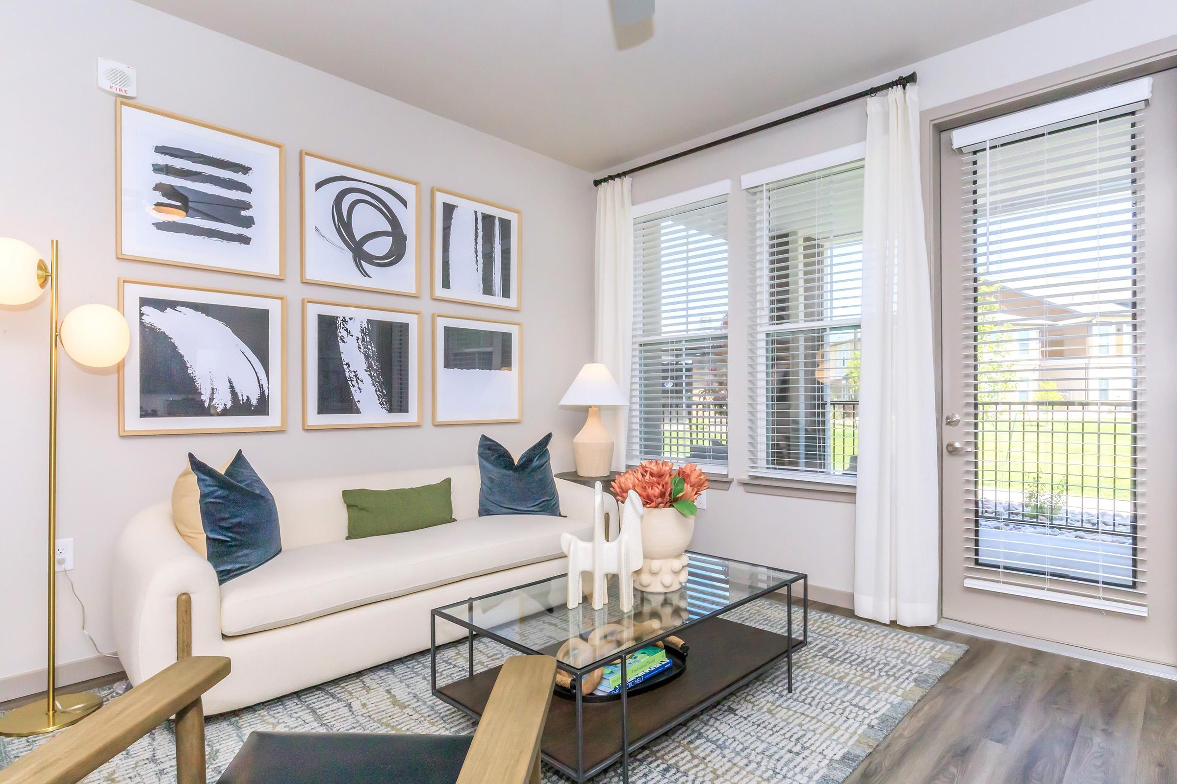 A modern living room featuring a white sofa with blue and green decorative pillows, a glass coffee table, and a vase of tulips. The walls are adorned with abstract artwork in frames. Natural light streams in through large windows with blinds, creating a bright atmosphere.